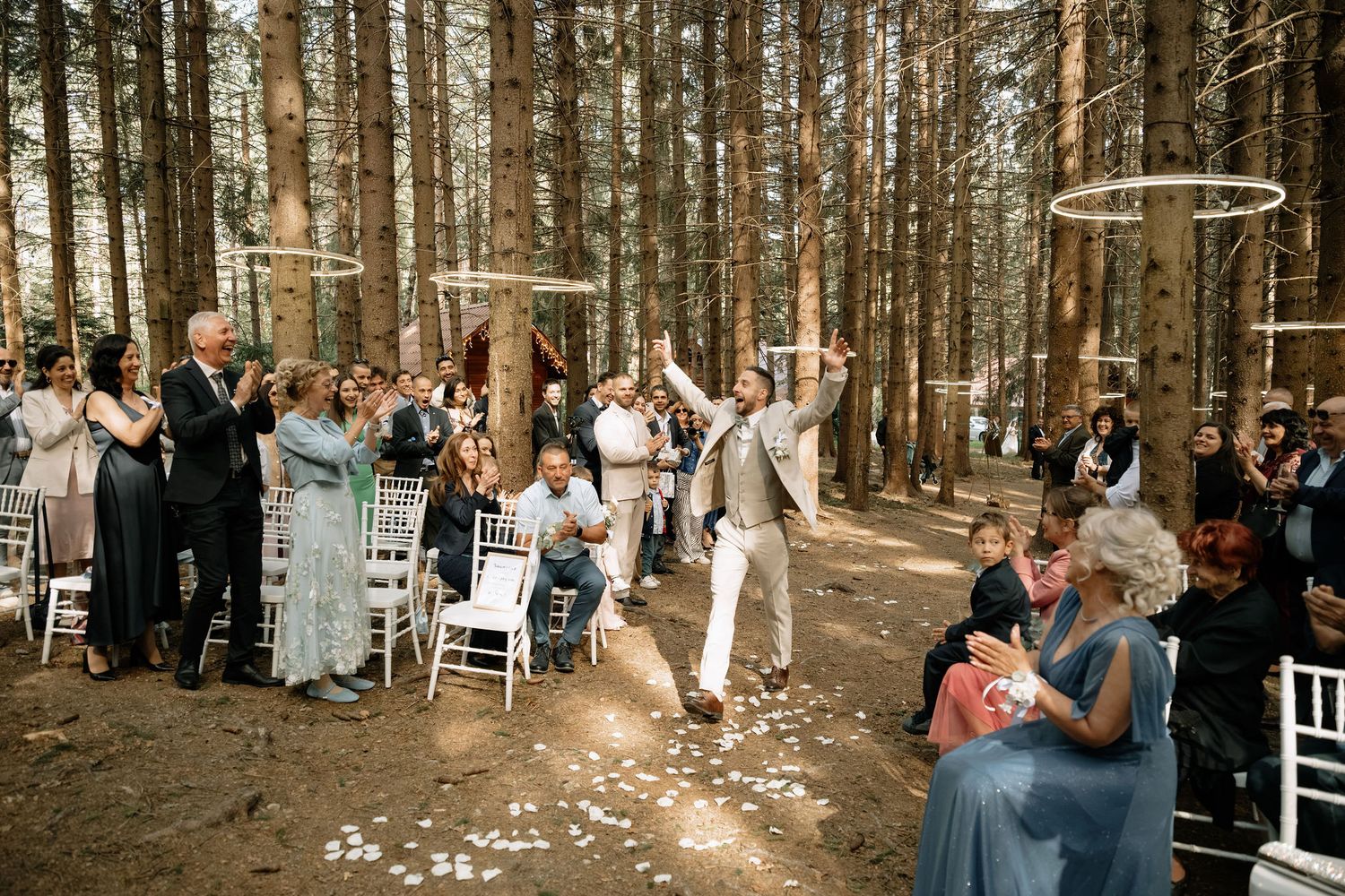 Joyful couple celebrates wedding ceremony in a sunlit pine forest with guests cheering along a white chair-lined aisle.