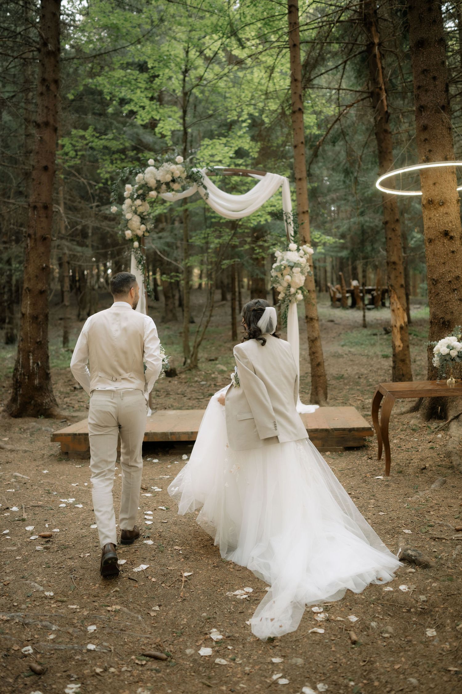 Bride and groom walking toward floral arch in forest wedding ceremony, viewed from behind.