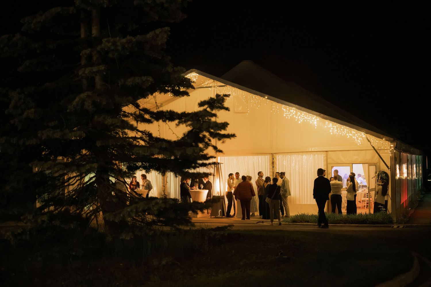 Elegant outdoor event tent glowing at night with guests mingling outside under string lights.