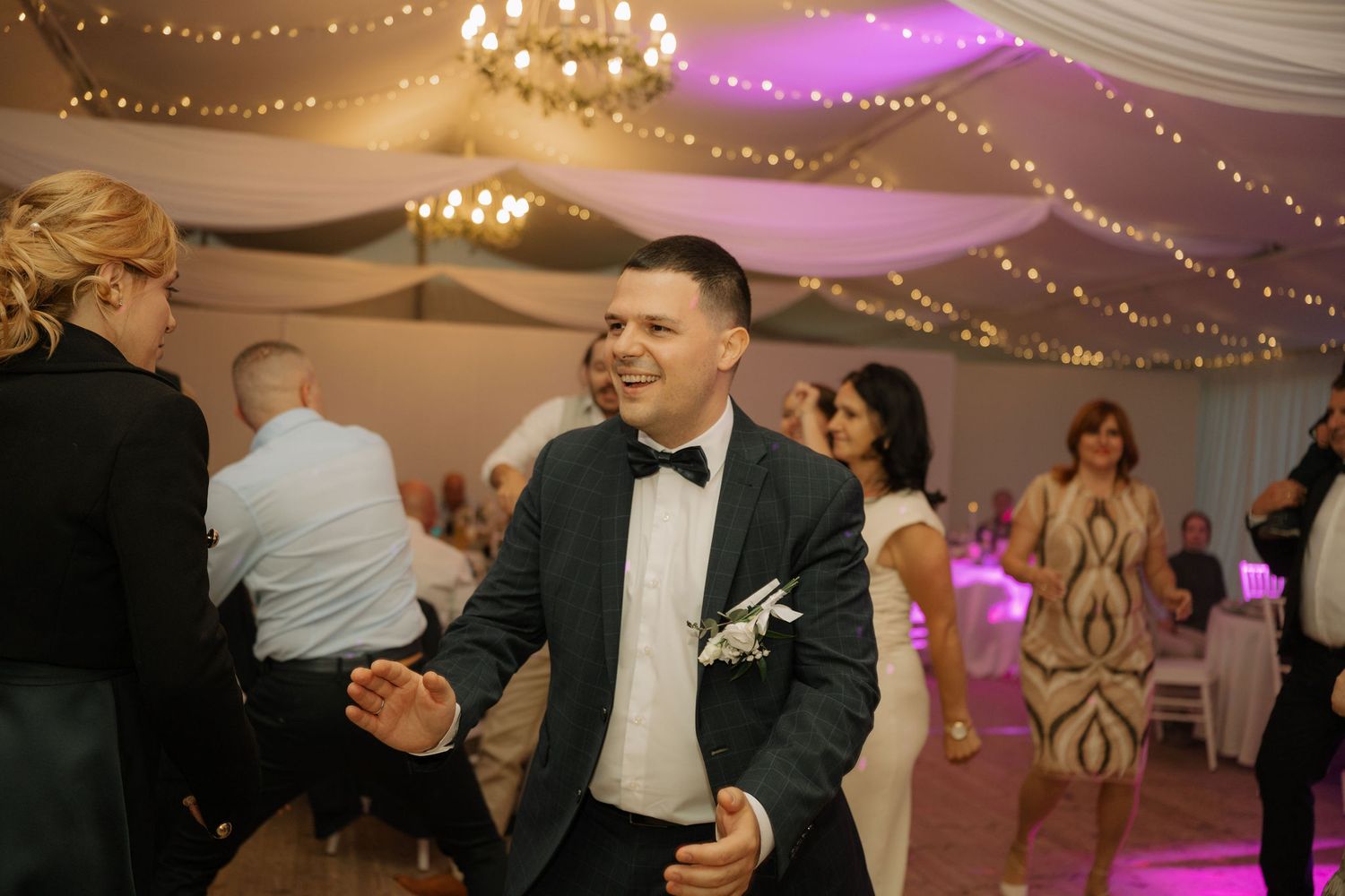 Groom in navy suit and bow tie dancing joyfully at elegant wedding reception with fairy lights and chandeliers.