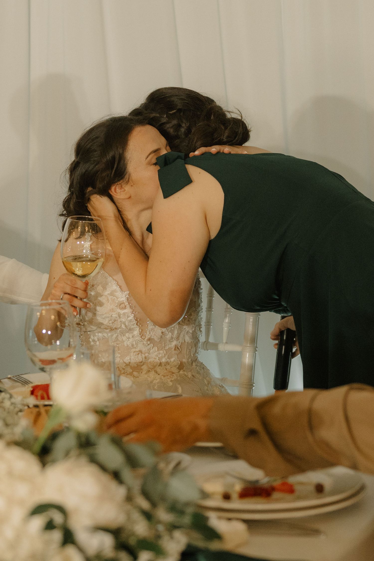 Bride in lace gown receiving a kiss on the cheek at wedding reception table with white flowers and wine glasses.