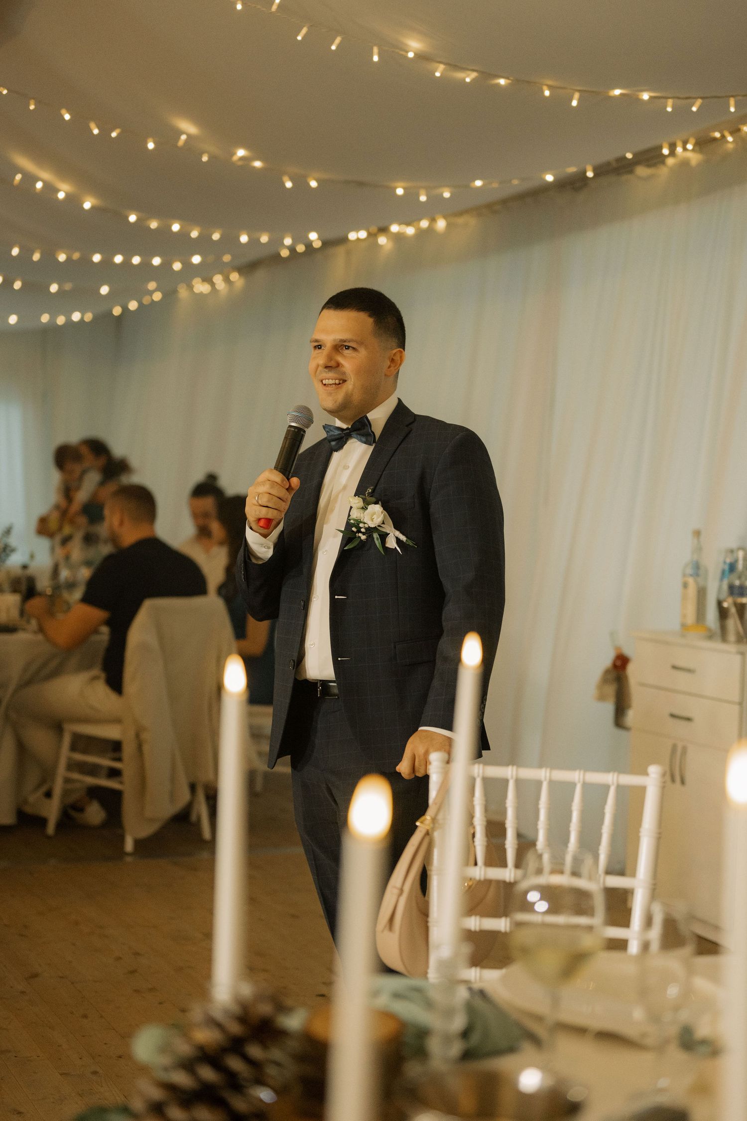 Smiling groom in navy suit with bow tie and boutonniere gives a speech at an elegant wedding reception with fairy lights.