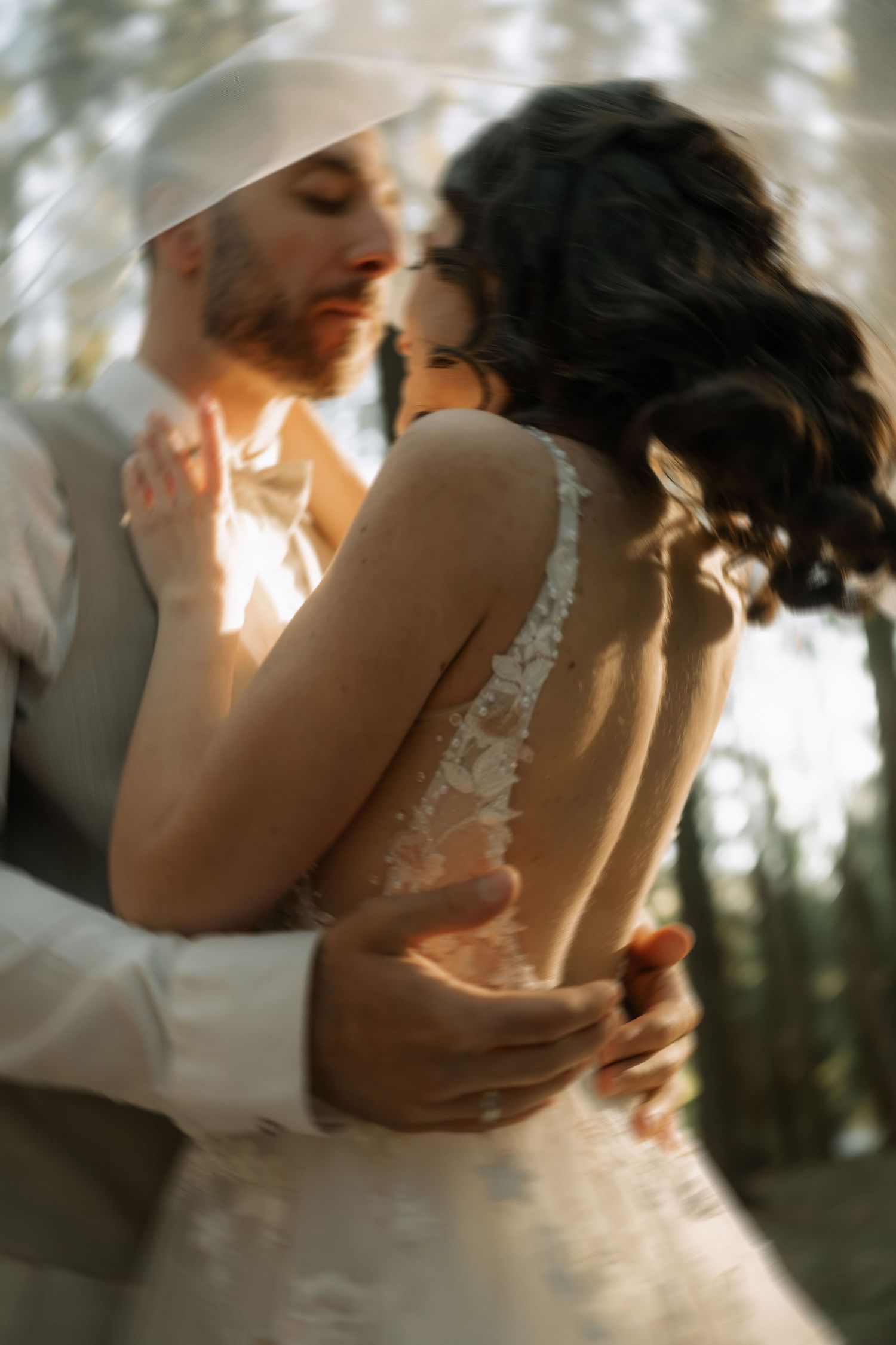 A bride and groom share an intimate moment under a veil, with soft golden light filtering through trees in a romantic outdoor setting.