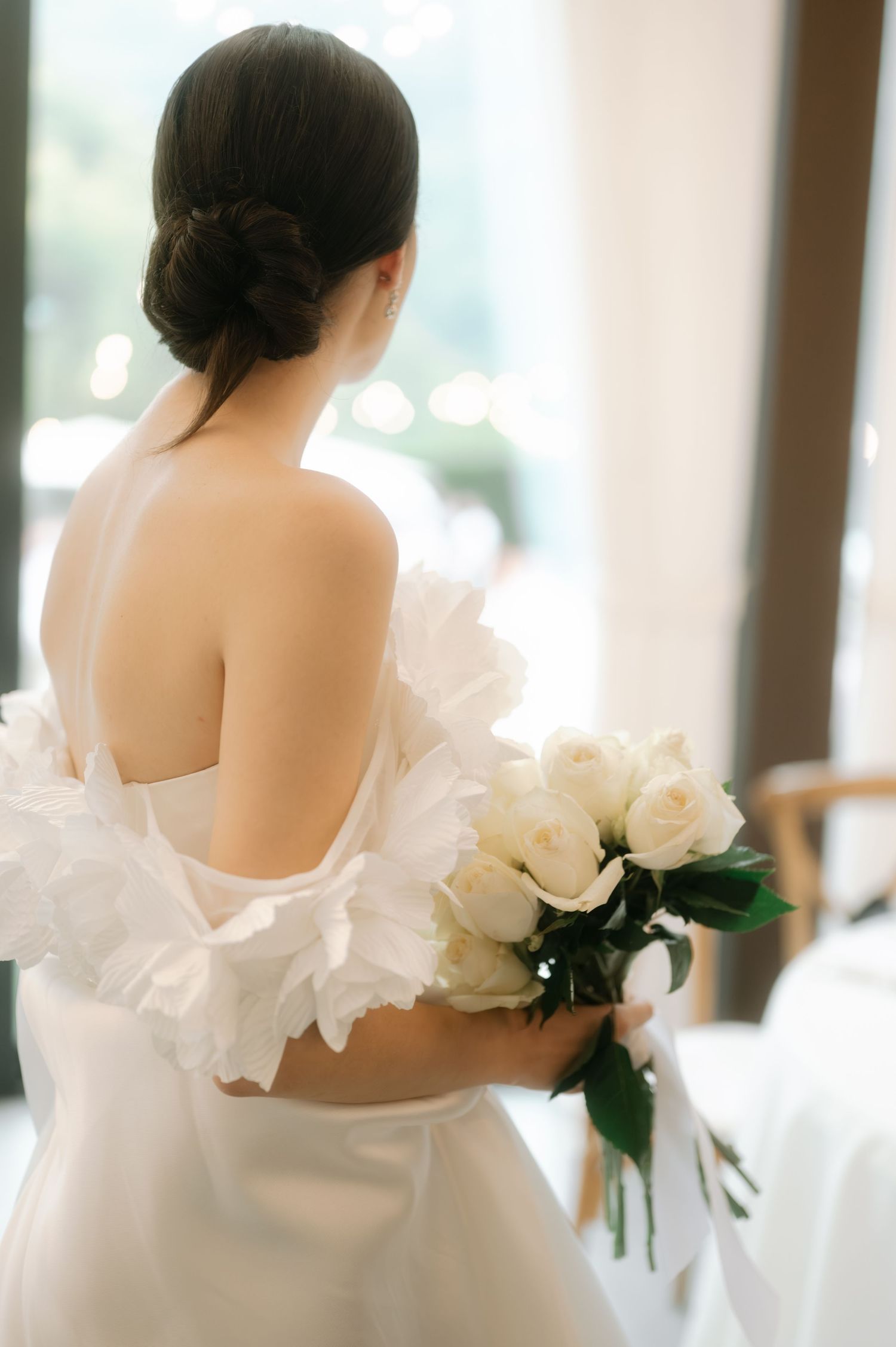 Three elegant white wedding dresses shown from behind with delicate ruffles and white peonies bouquets in a bright room.