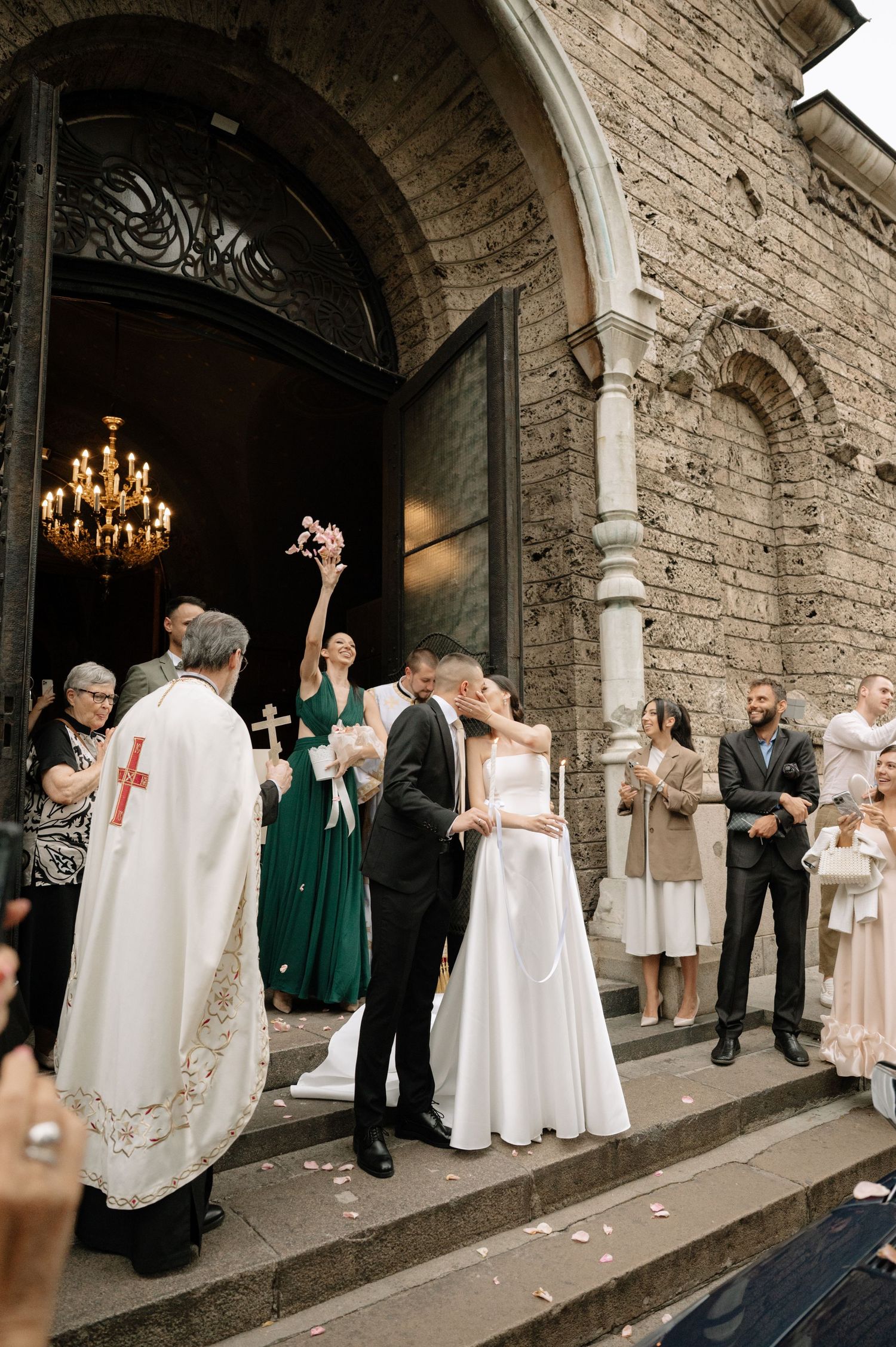 A traditional church wedding ceremony takes place at the entrance of a historic stone church with a chandelier visible inside.