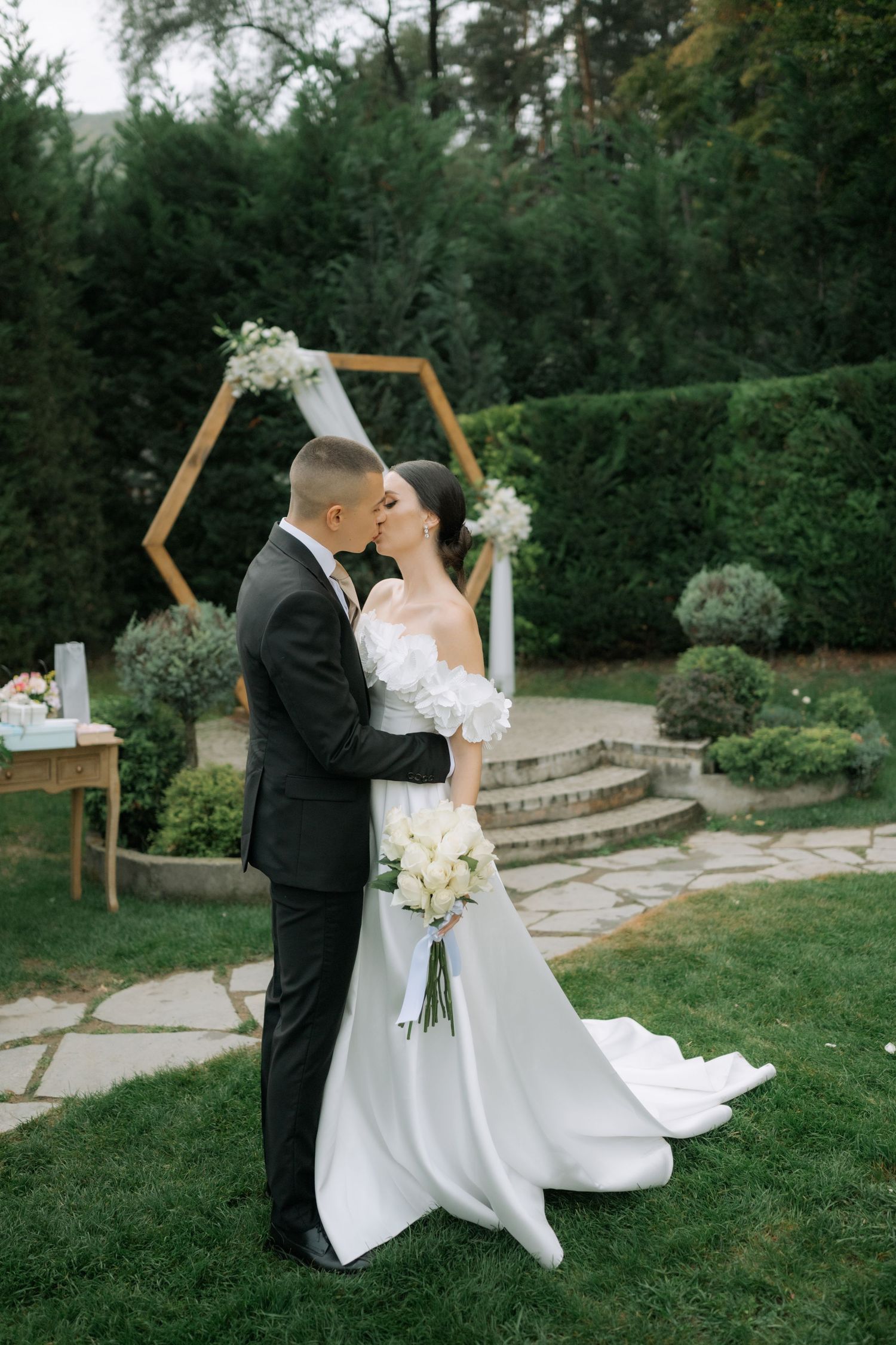 Romantic wedding couple sharing a tender kiss under a wooden hexagonal arch in a lush garden setting.