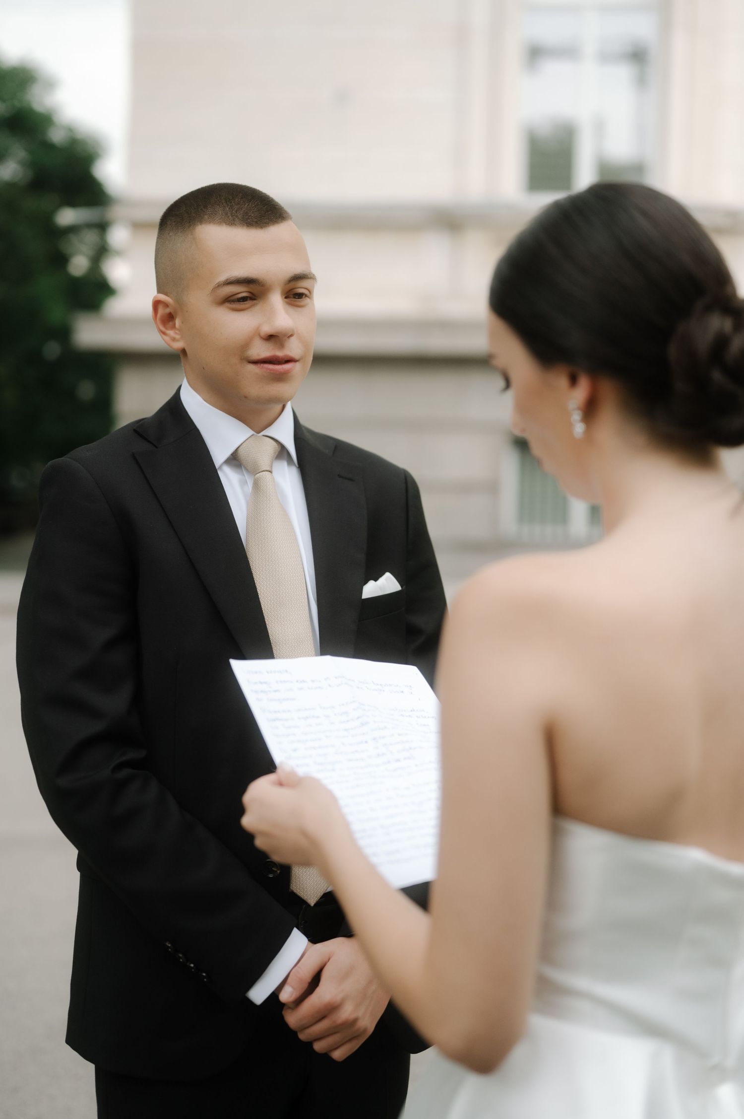 Couple in wedding attire exchanging vows while reading from papers during outdoor ceremony.