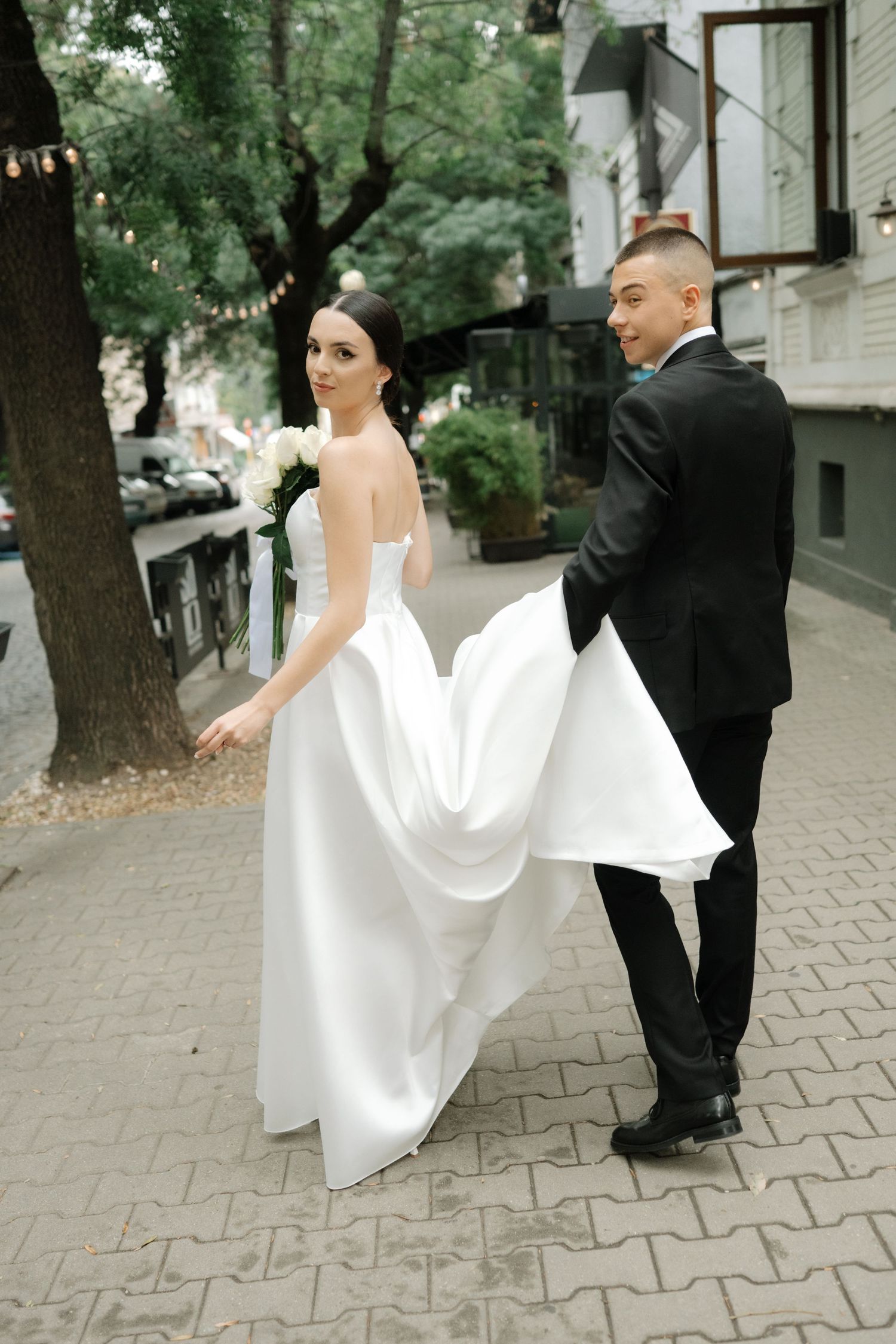 A couple on their wedding day standing on a city sidewalk, with the bride in white and groom in black tuxedo.