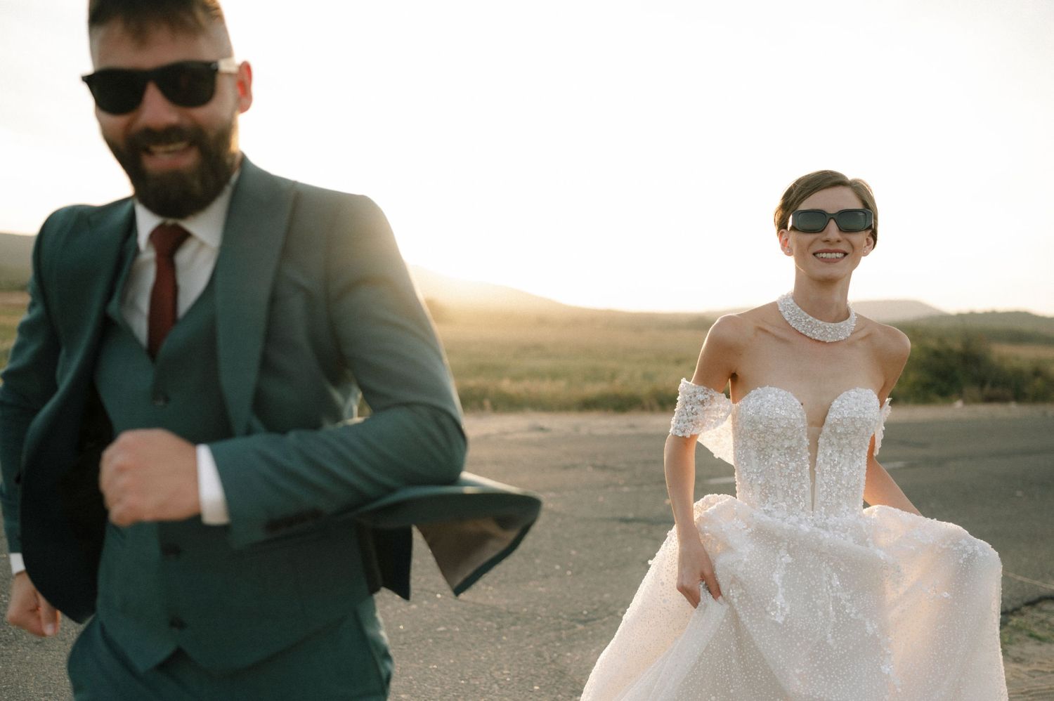 Bride in white gown and groom in teal suit pose outdoors at sunset during a stylish wedding photo shoot.