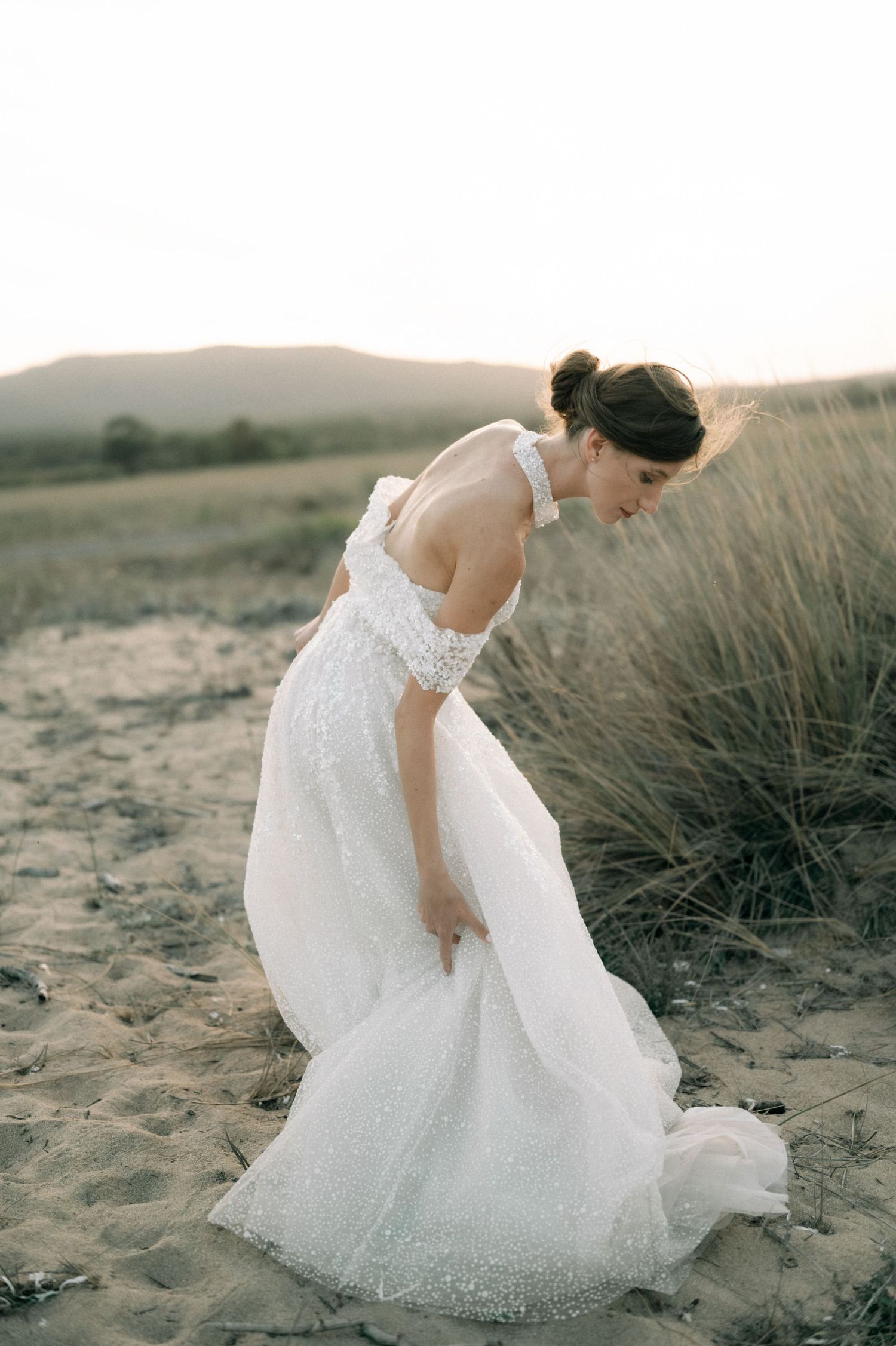 A bride in a white sequined off-shoulder gown bends gracefully on sandy dunes at sunset.