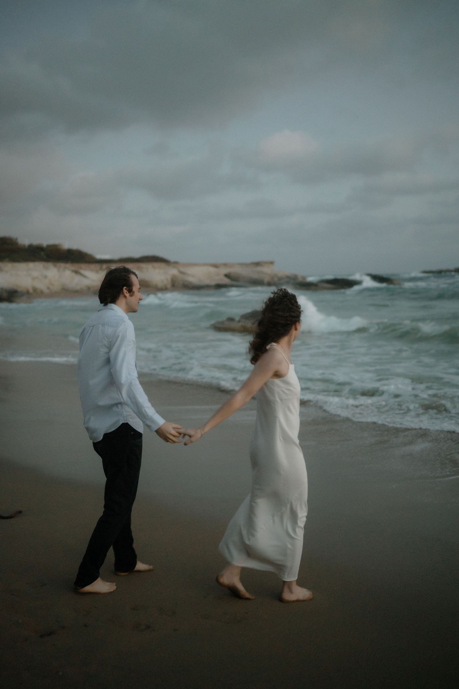 Couple holding hands walking barefoot on a moody beach with waves and rocky cliffs in the background.