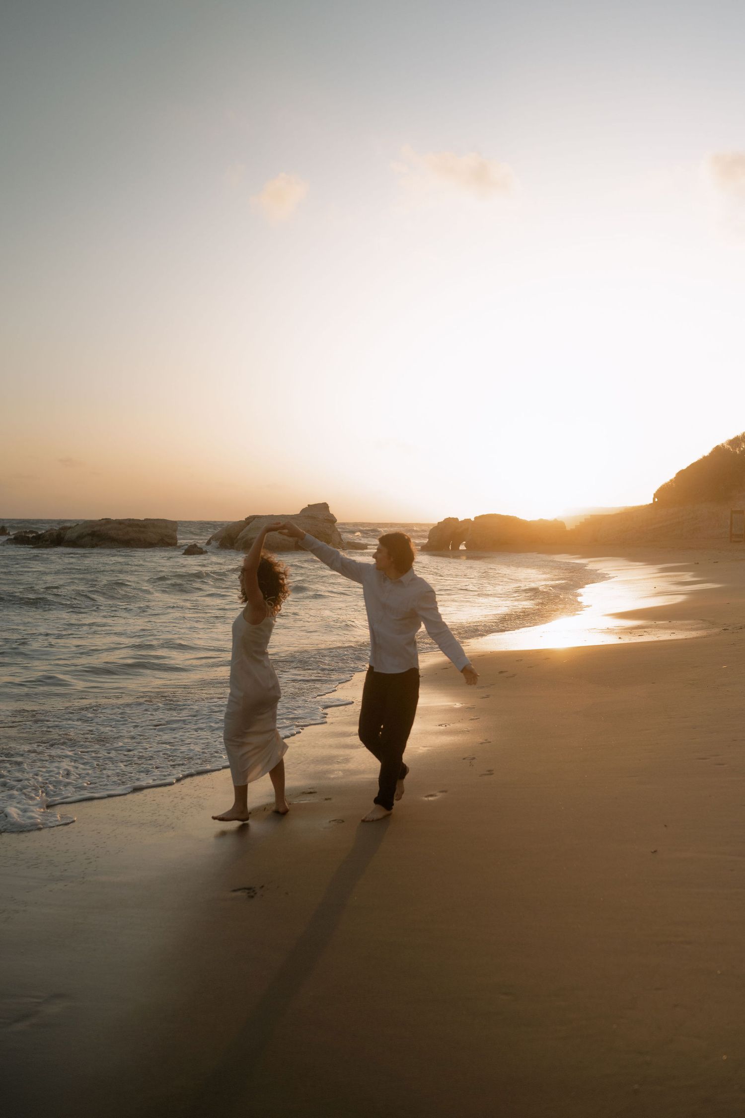 Couple dancing on a sandy beach at golden sunset, waves gently lapping the shore beside rocky cliffs.
