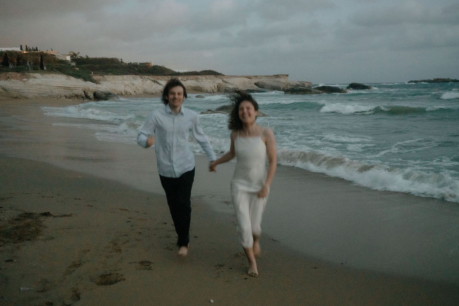 Couple holding hands running barefoot on a sandy beach at dusk with ocean waves in the background.