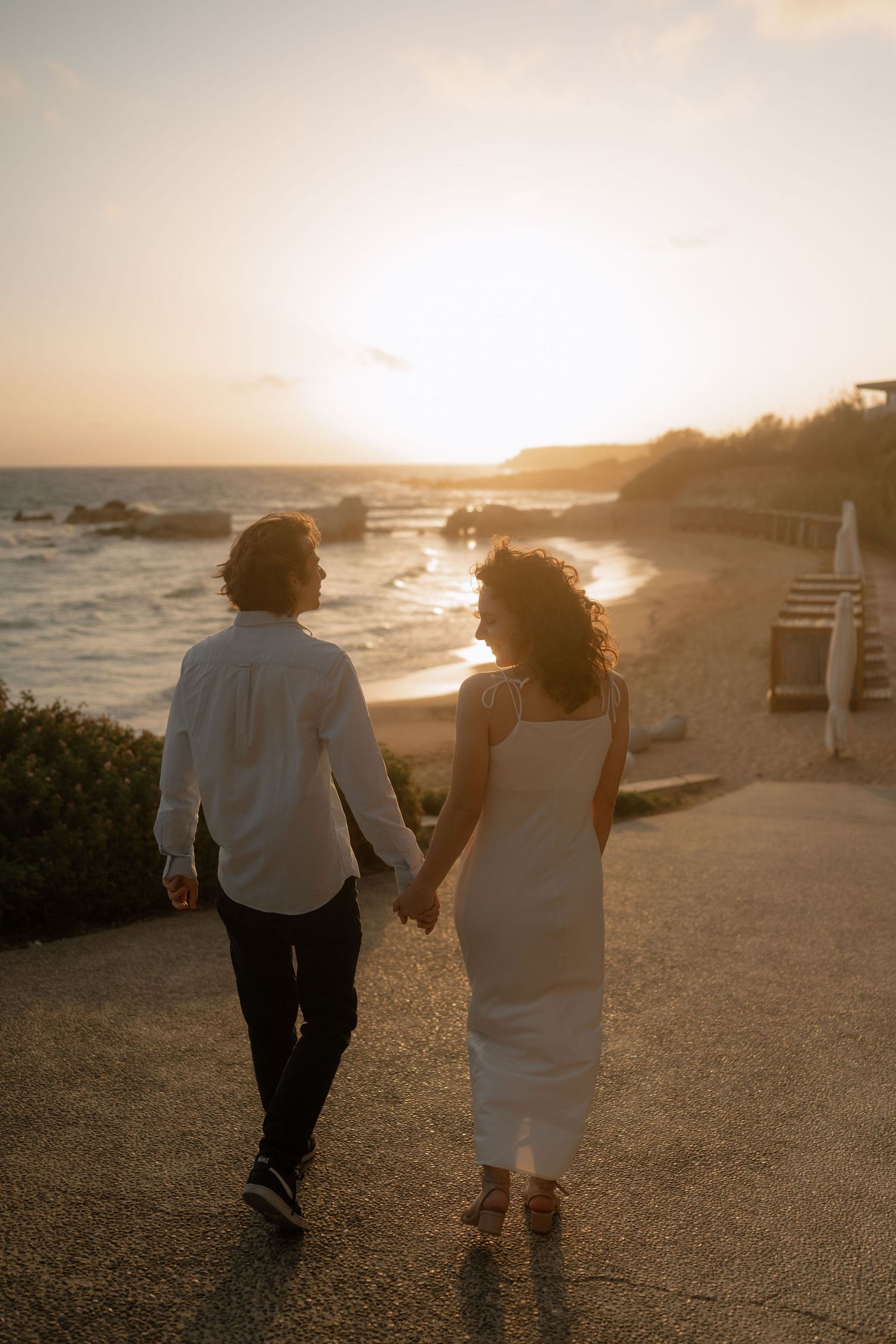 Couple holding hands walking toward a stunning ocean sunset on a coastal path, woman in white dress.