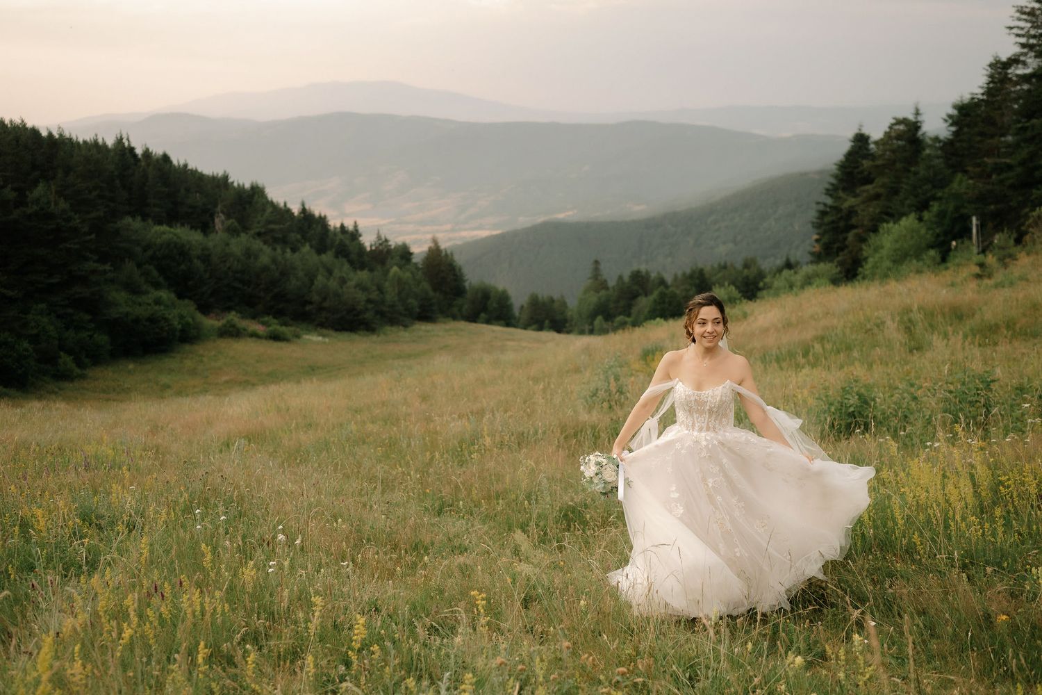A scenic mountain meadow at sunset with rolling hills and evergreen forests in the background.