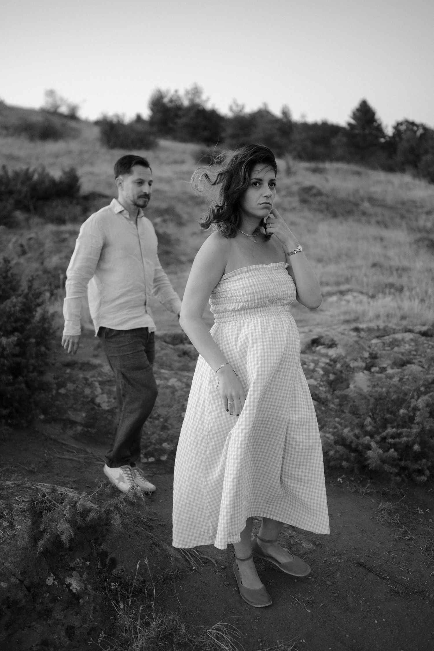 A couple in formal attire dances playfully together on a hillside in black and white photography.