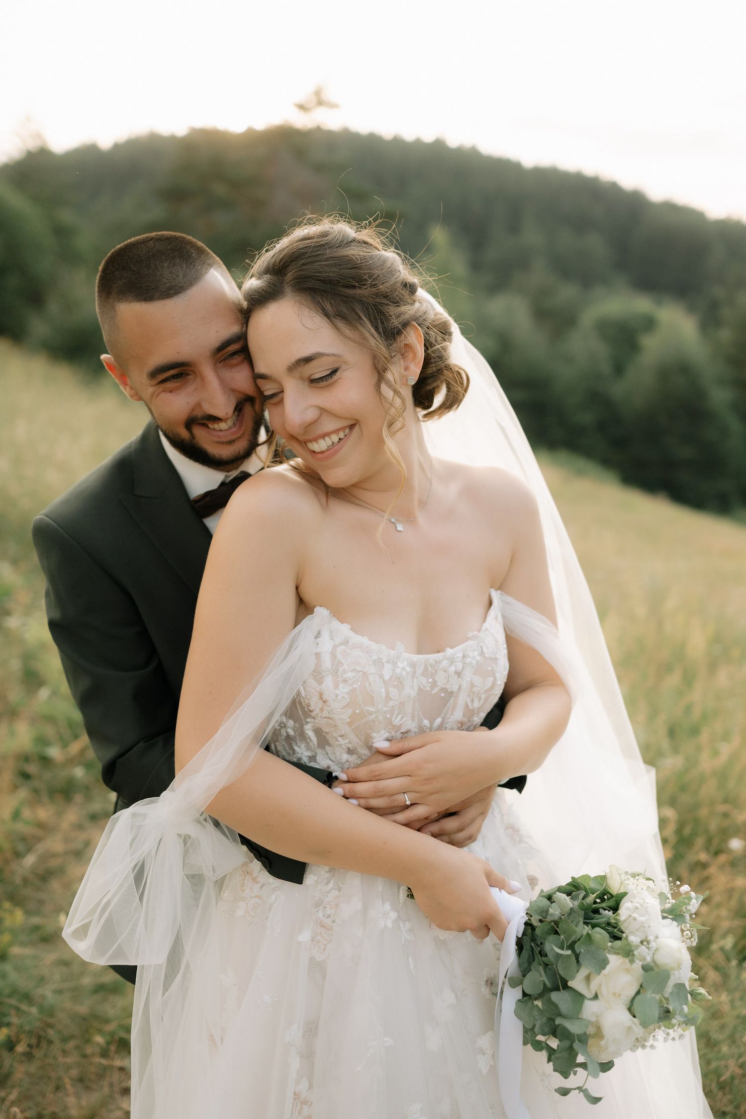 A romantic wedding moment captured outdoors with a couple embracing in a sunlit meadow with mountains in the background.