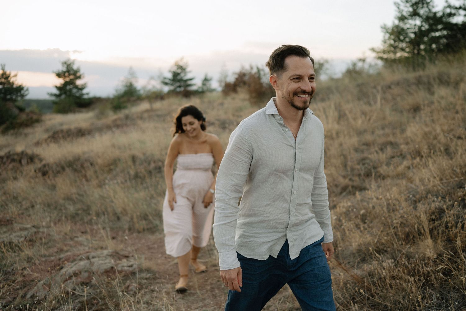 A couple shares an intimate moment while walking through tall grass on a hillside at sunset.