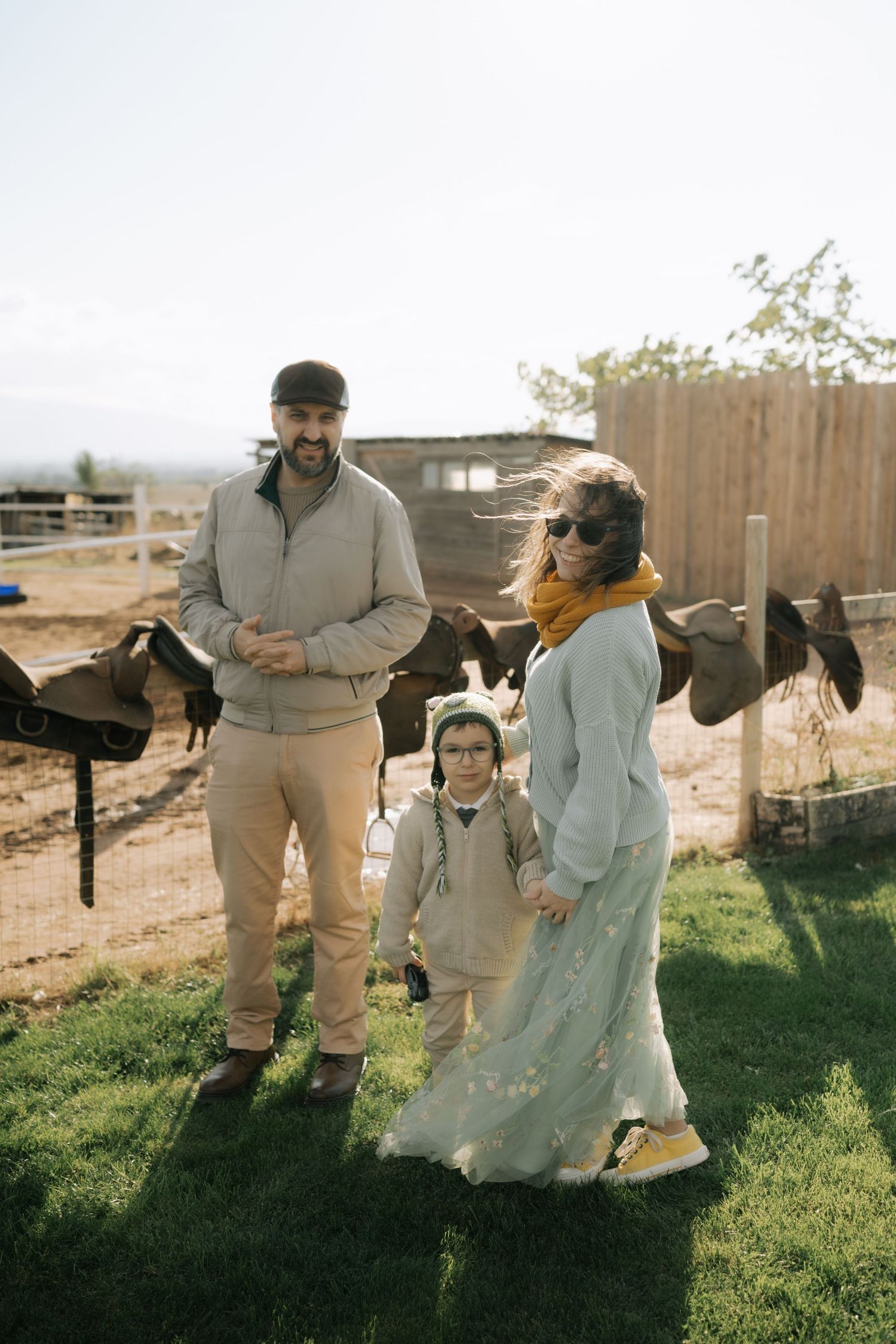A family stands together on a grassy hill near a wooden fence with horses in the background on a sunny day.