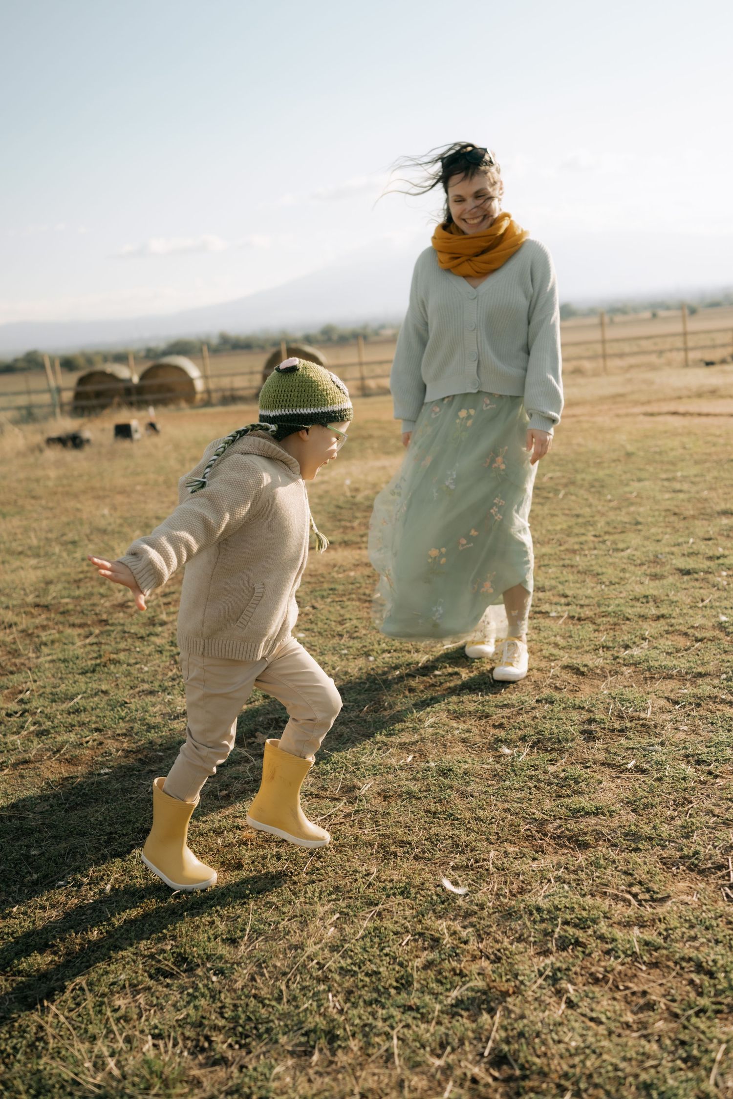 Two people in casual attire playfully chase each other across a grassy field on a sunny afternoon.