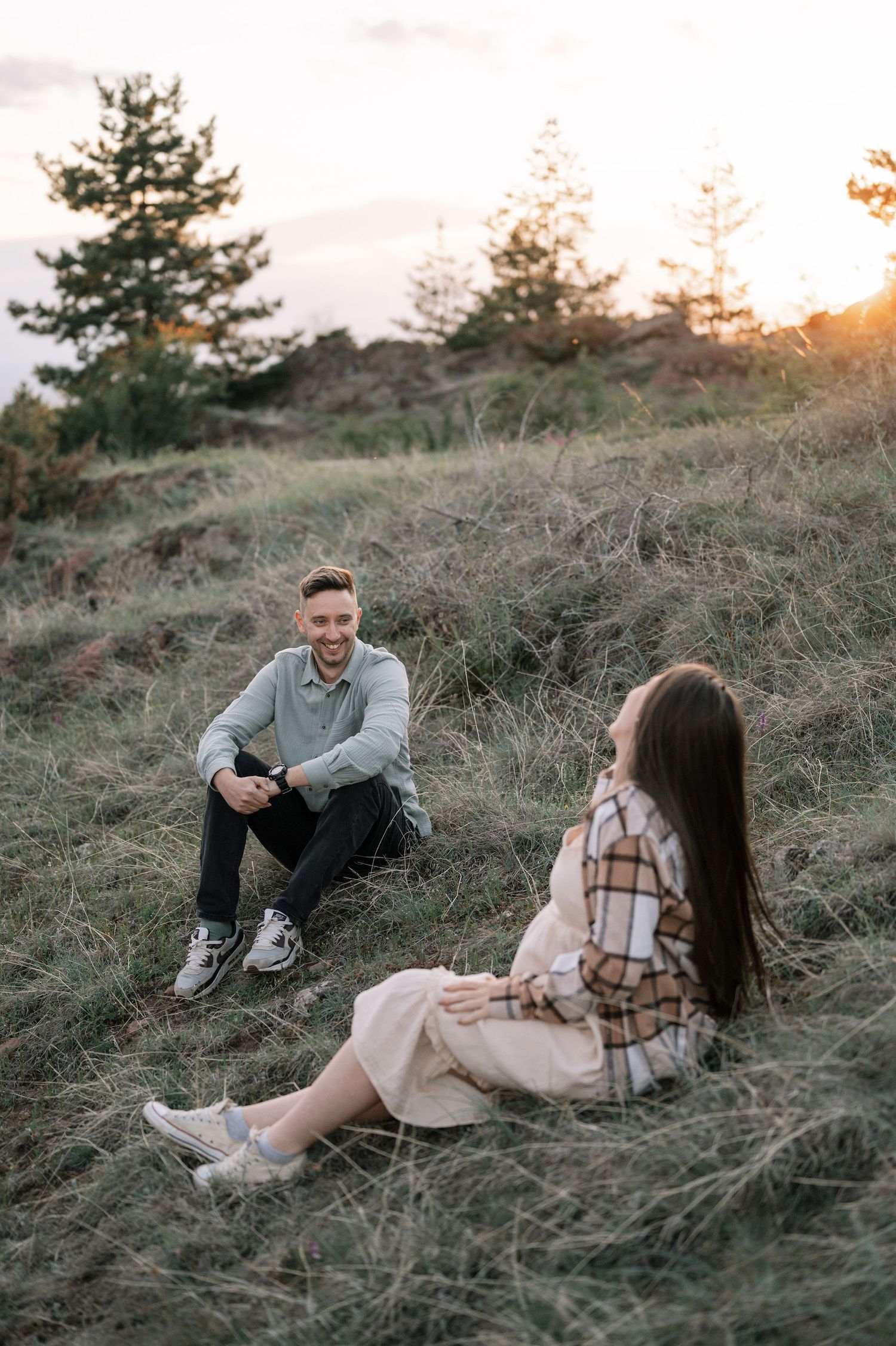 A couple sits together on a grassy hillside at sunset sharing an intimate moment in casual attire.