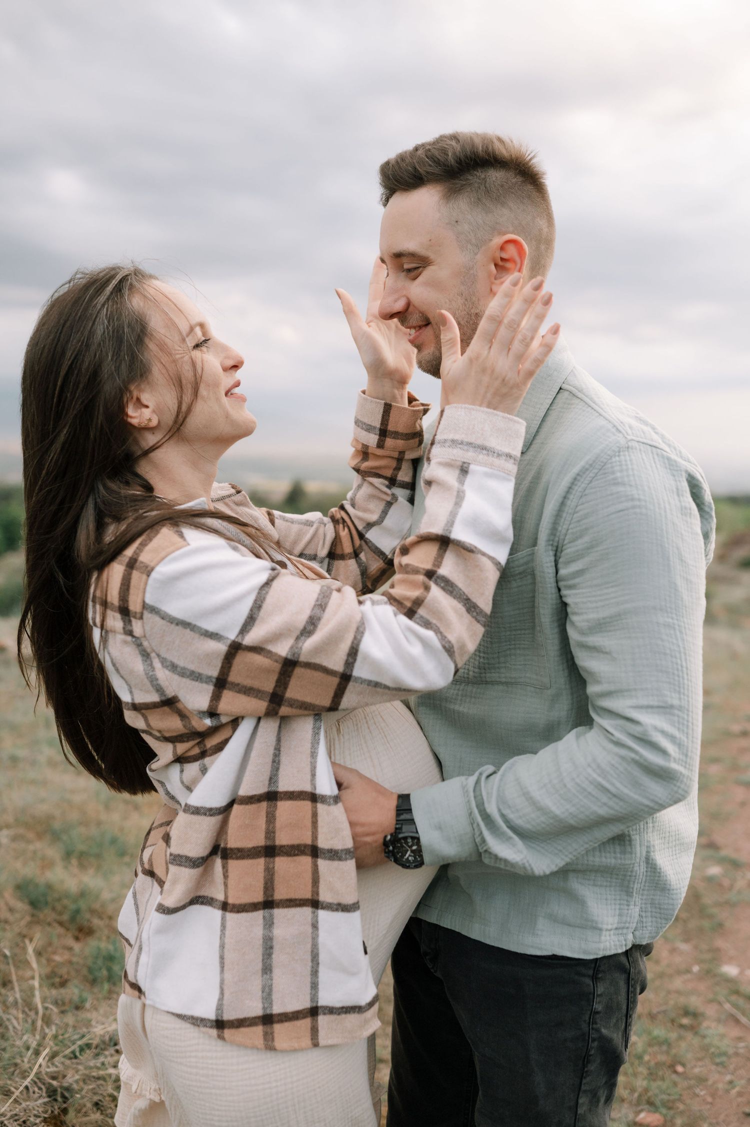 A couple shares a joyful embrace in a field on a cloudy day, wearing cozy sweaters and a plaid scarf.