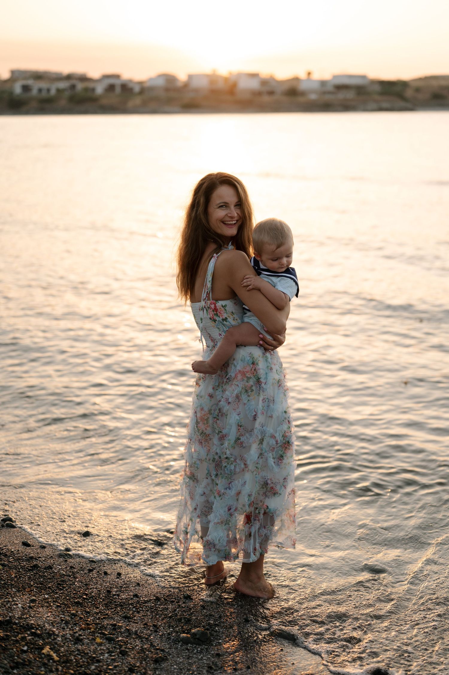 A parent and child share a tender moment at the beach during golden hour as gentle waves lap at their feet.