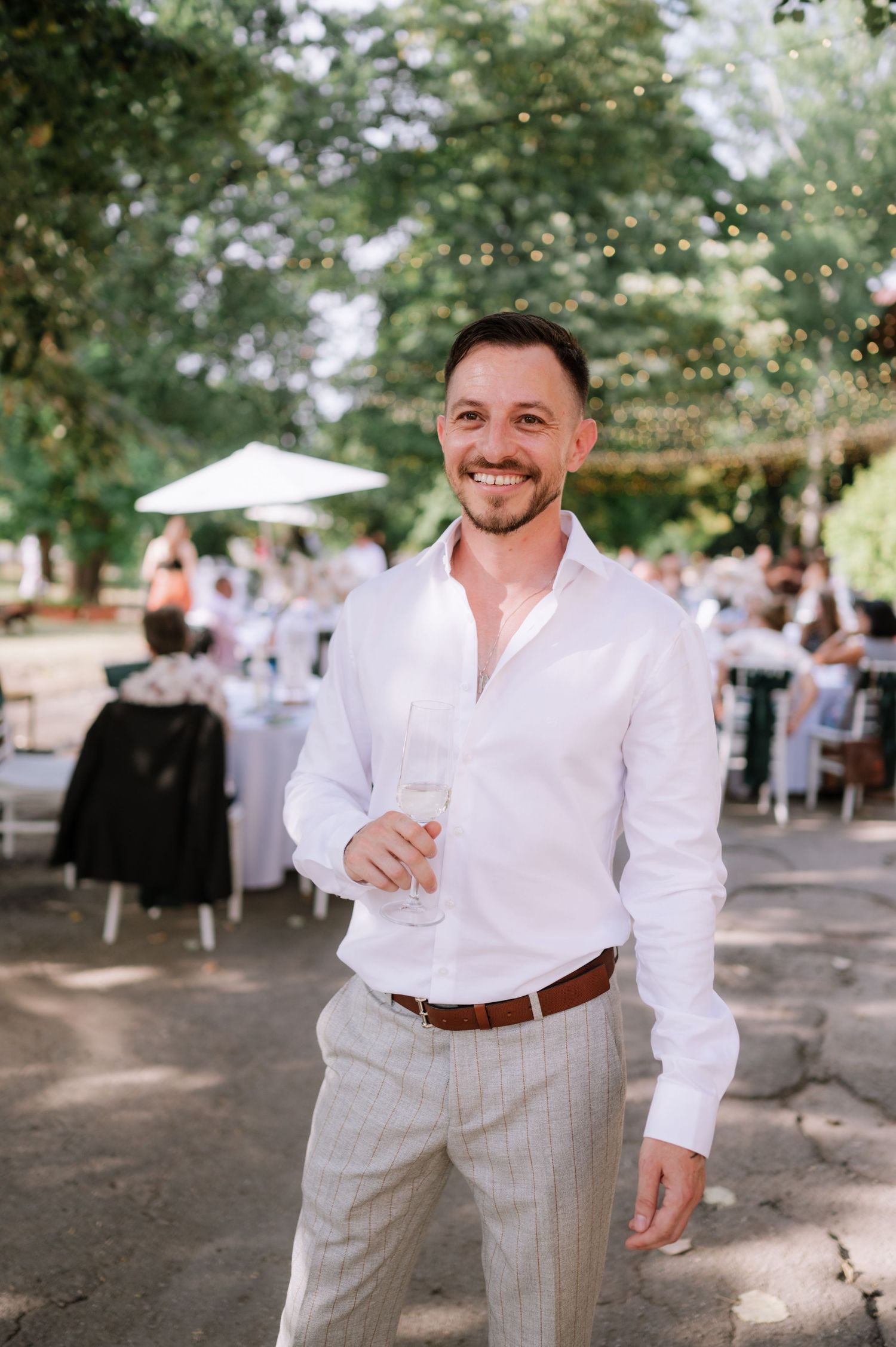 A person in a white button-down shirt and light gray pants stands outdoors at a casual summer event.