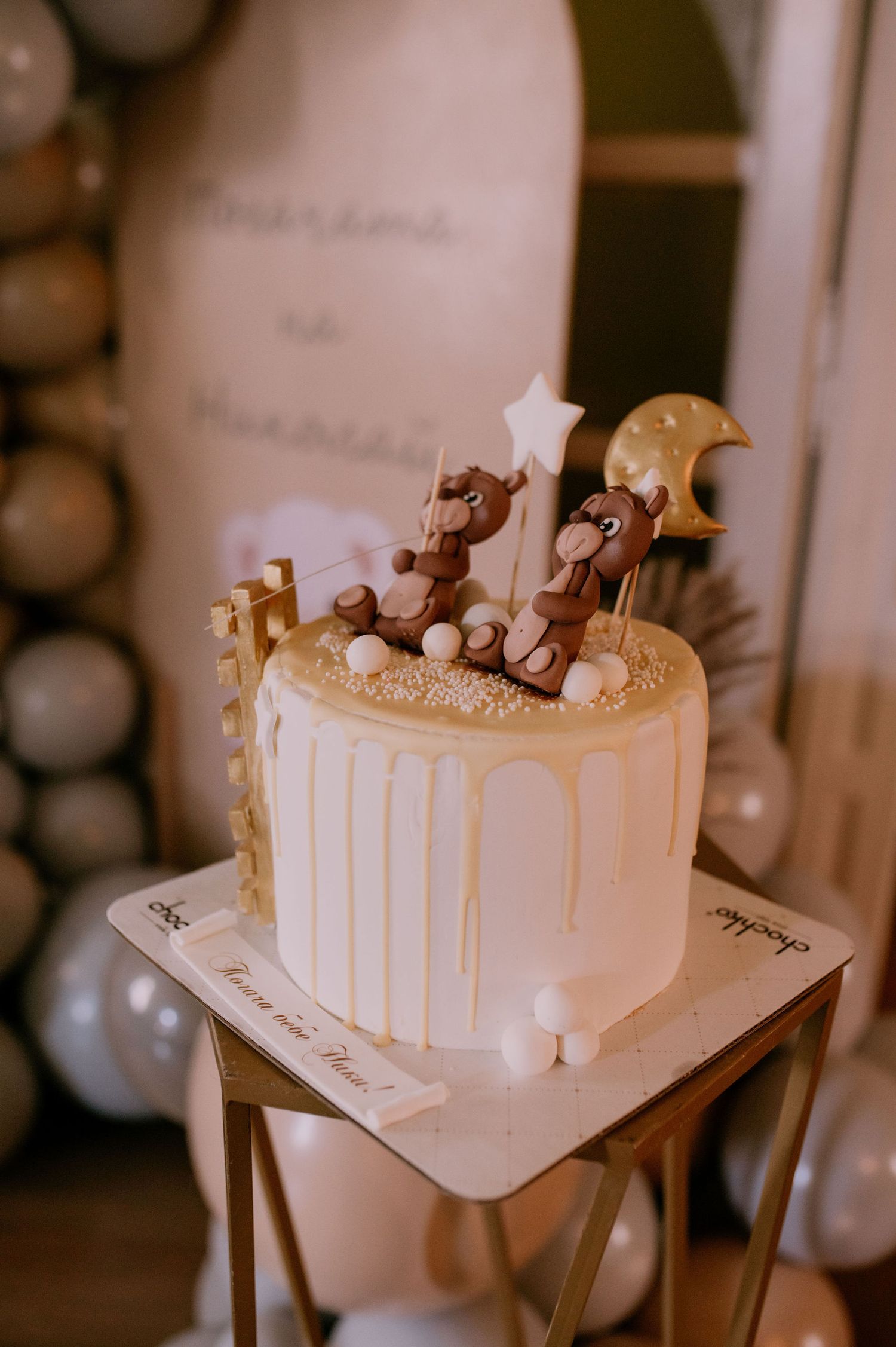 White wedding cake with drip details and decorative toppings displayed on wooden table in romantic lighting.