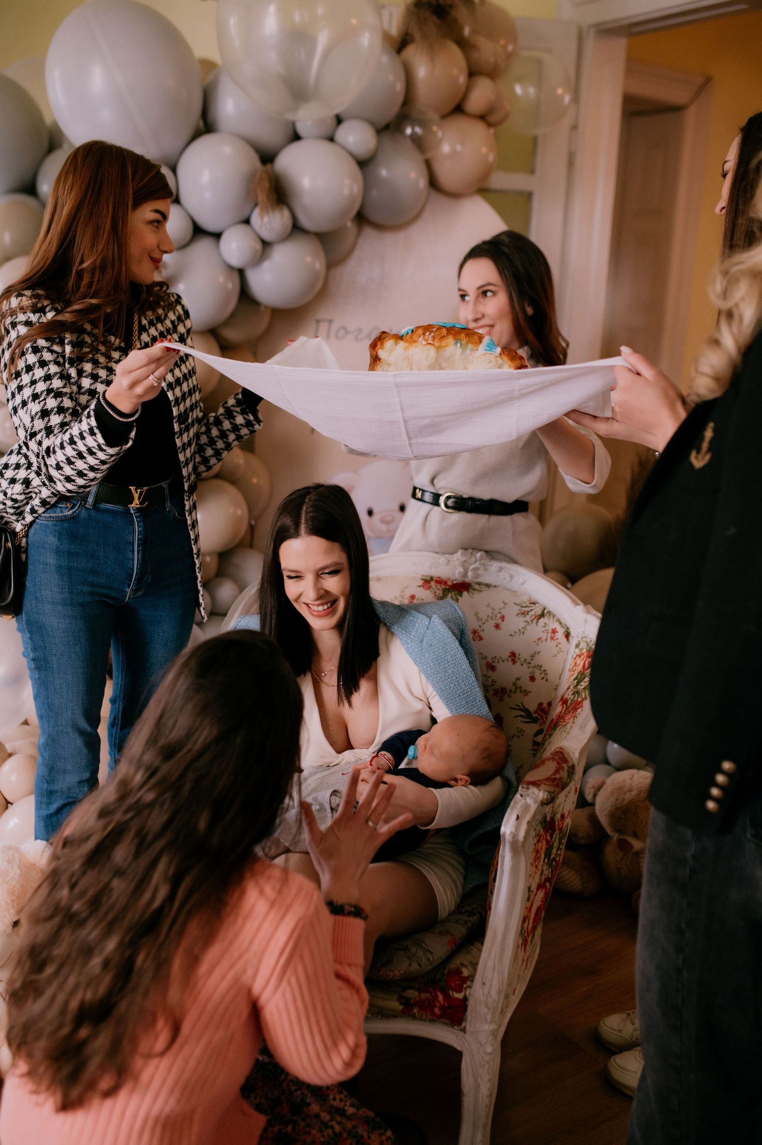 A group of friends gather at a party with balloon decorations, sharing laughter and conversation in a cozy indoor setting.