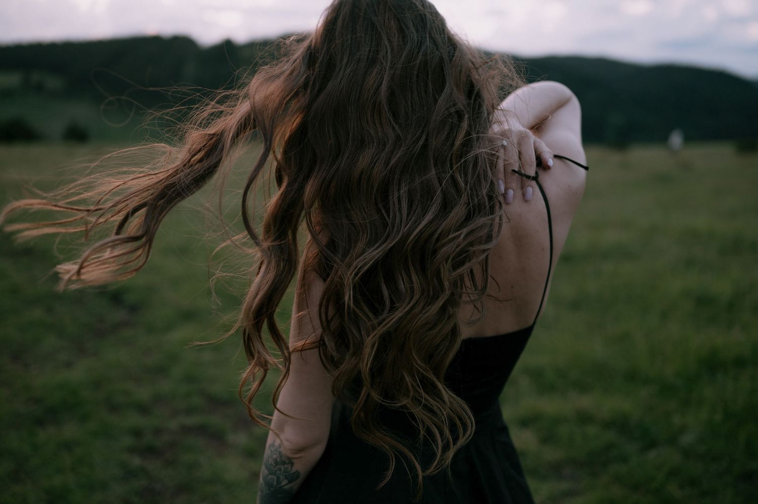 Long wavy brown hair blowing in the wind against a green countryside landscape on an overcast day.