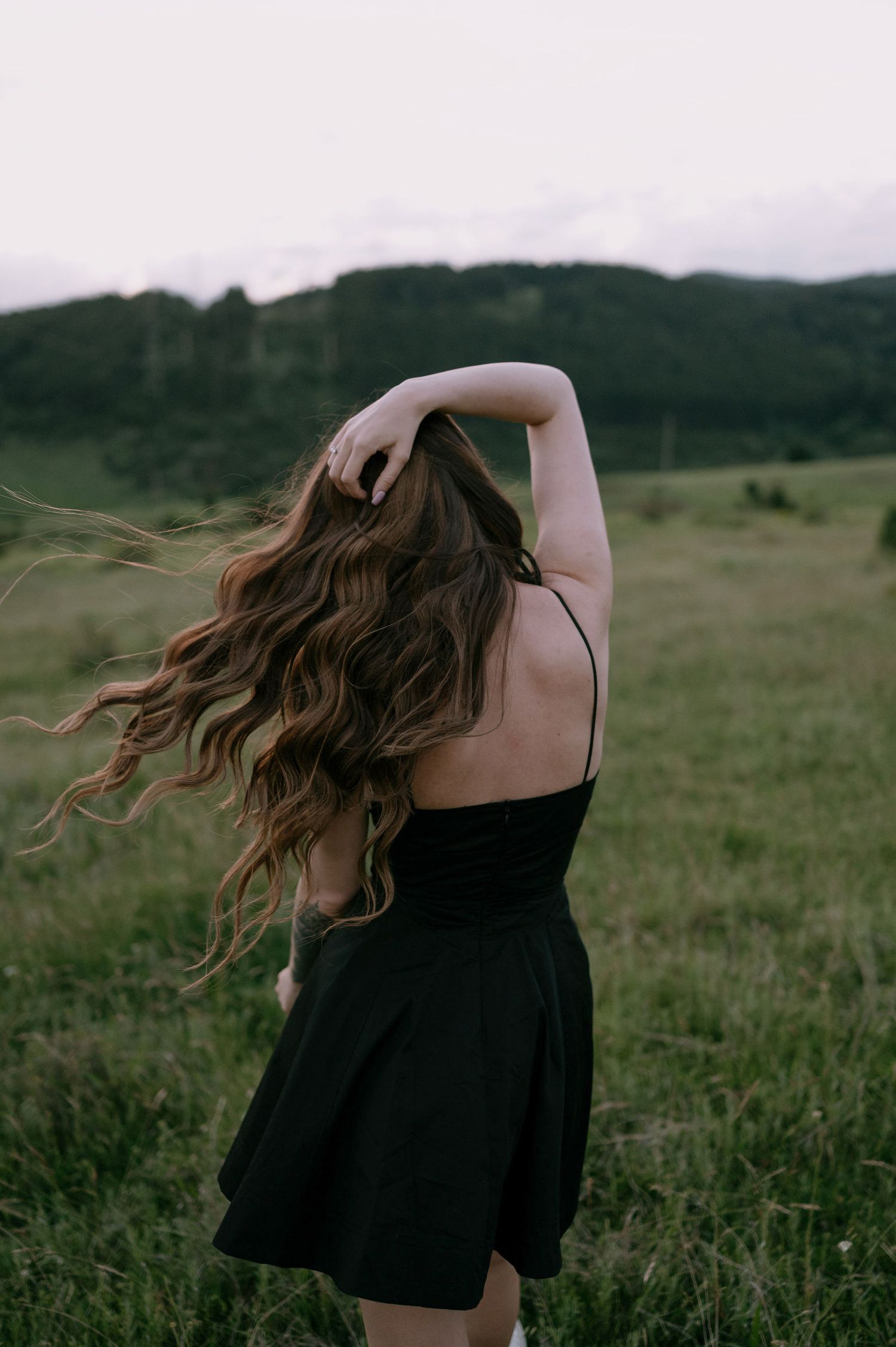 A person in a black dress with flowing long hair stands in a grassy field at dusk.