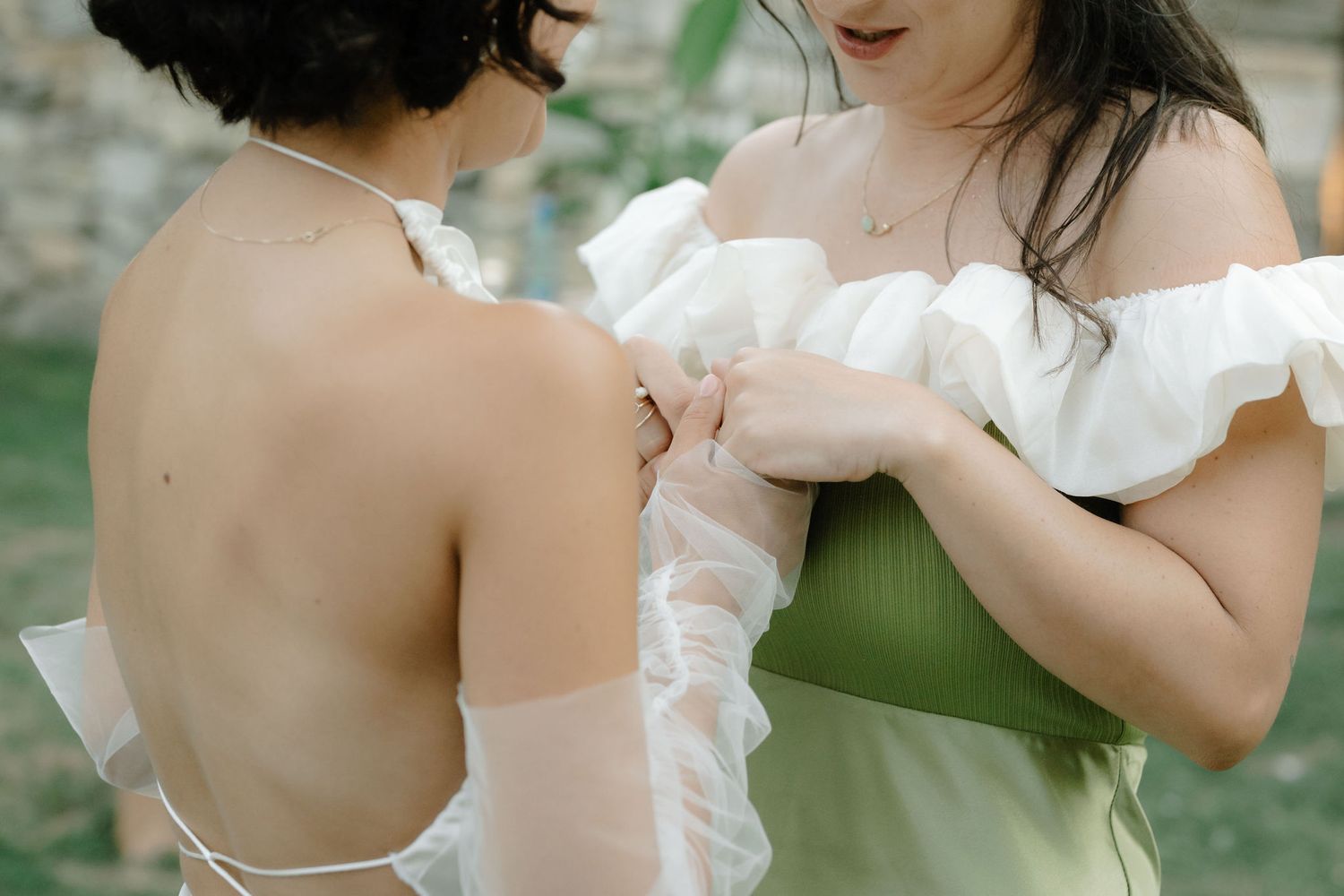 Two people wearing white off-shoulder dresses stand close together in an outdoor setting with greenery in background.