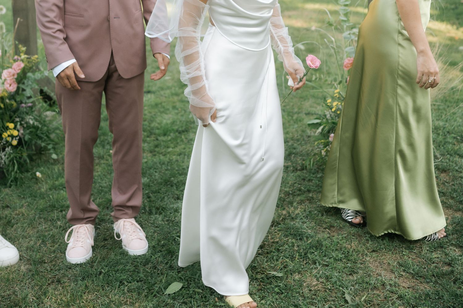 Wedding party members in brown suits and sage green dresses stand together on a grassy lawn during an outdoor celebration.