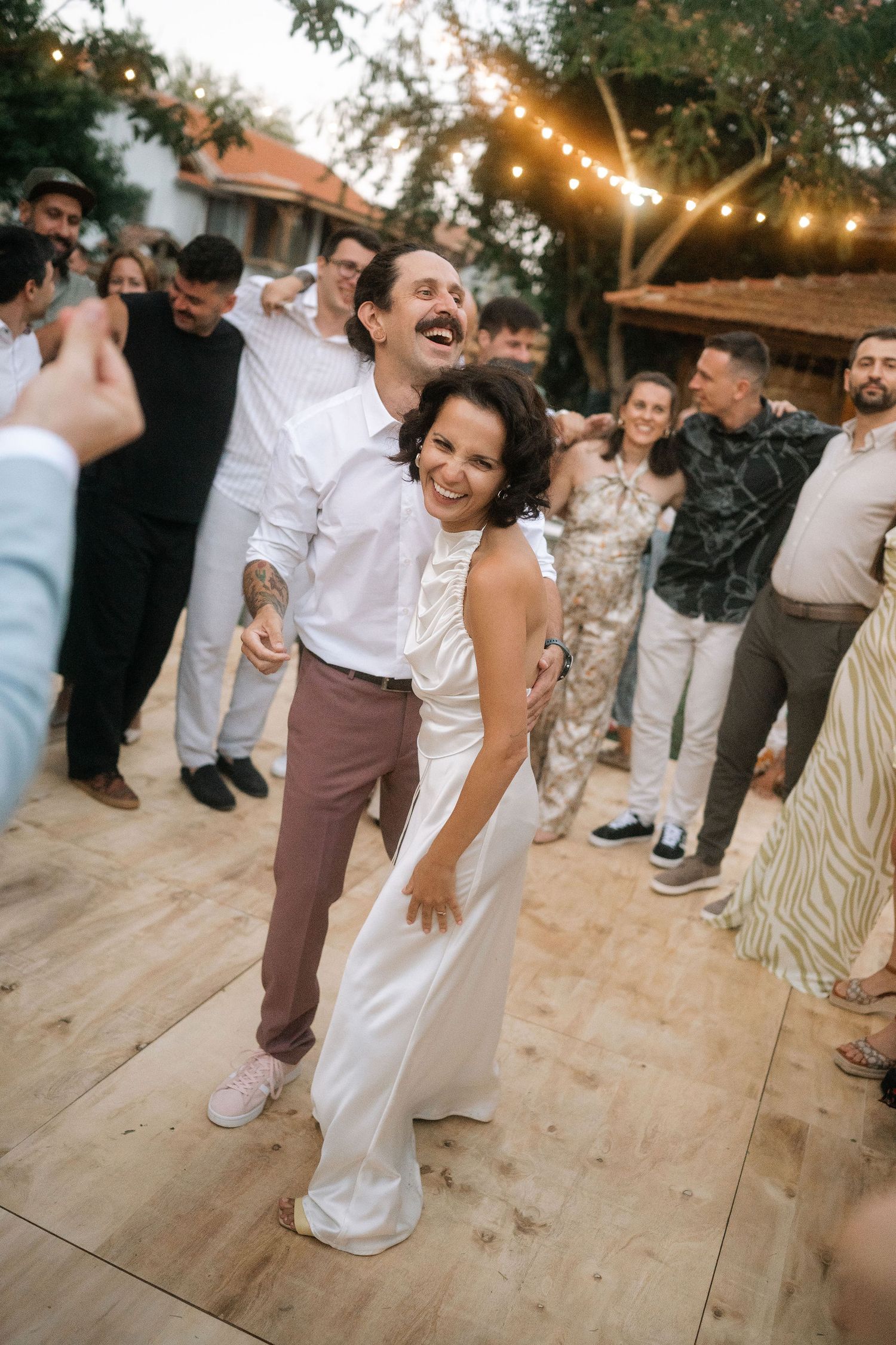 A couple laughs while dancing on a wooden deck at a sunset wedding celebration with guests watching.