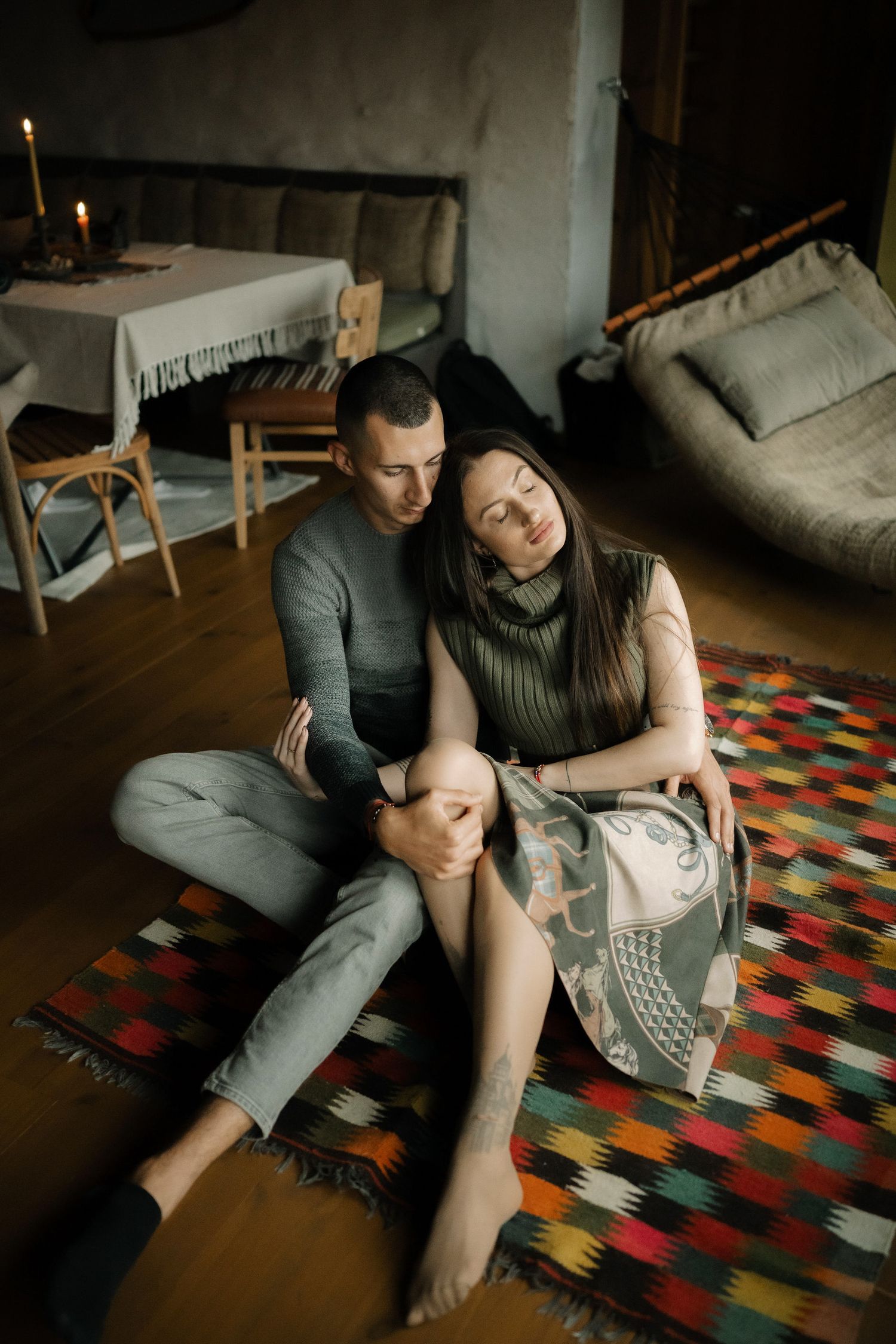 A couple relaxes together on a colorful patterned rug in a cozy room with candlelight and casual home decor.