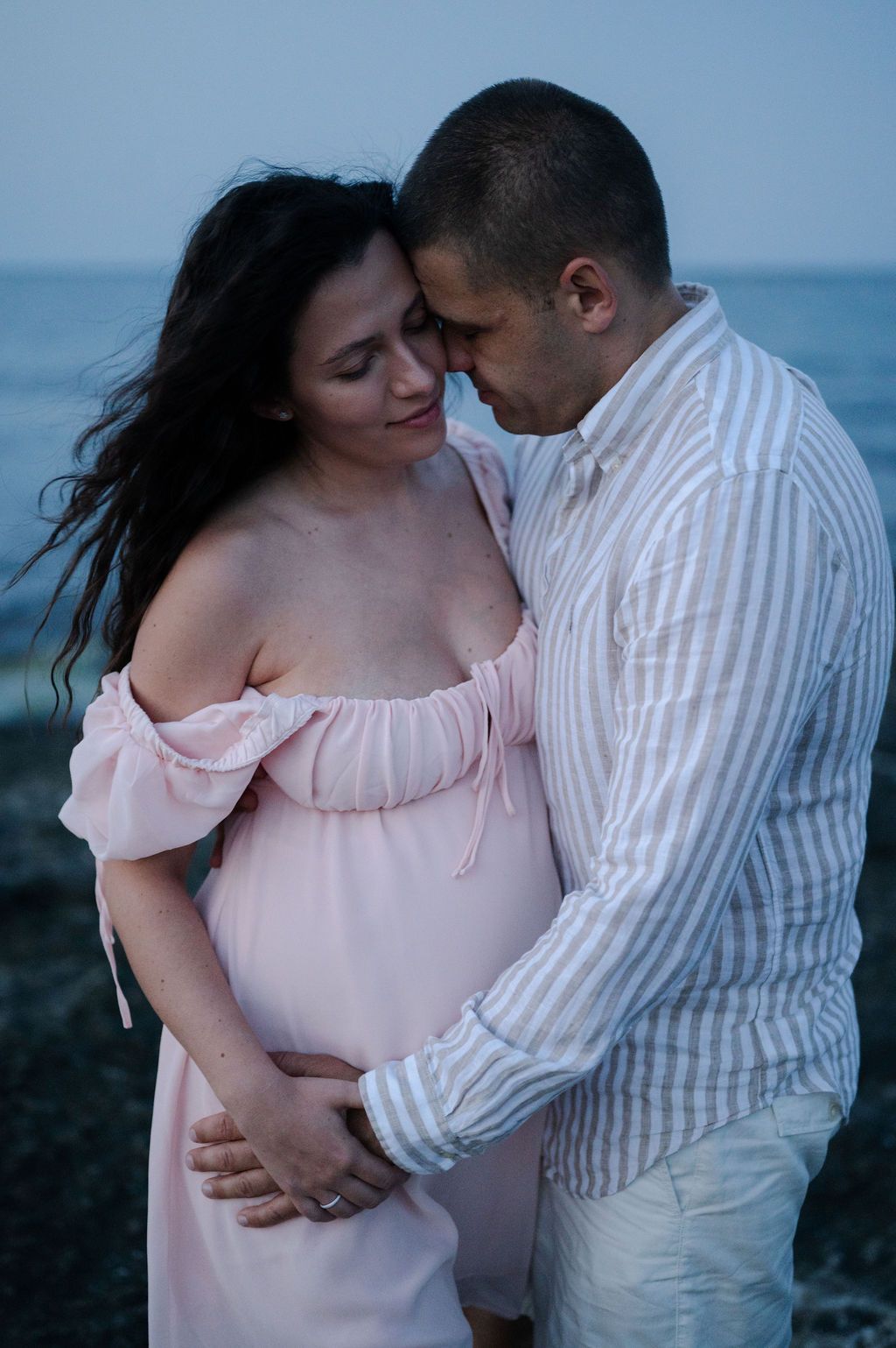 Romantic couple embraces at twilight by the ocean, wearing white and pink outfits in a series of intimate poses.