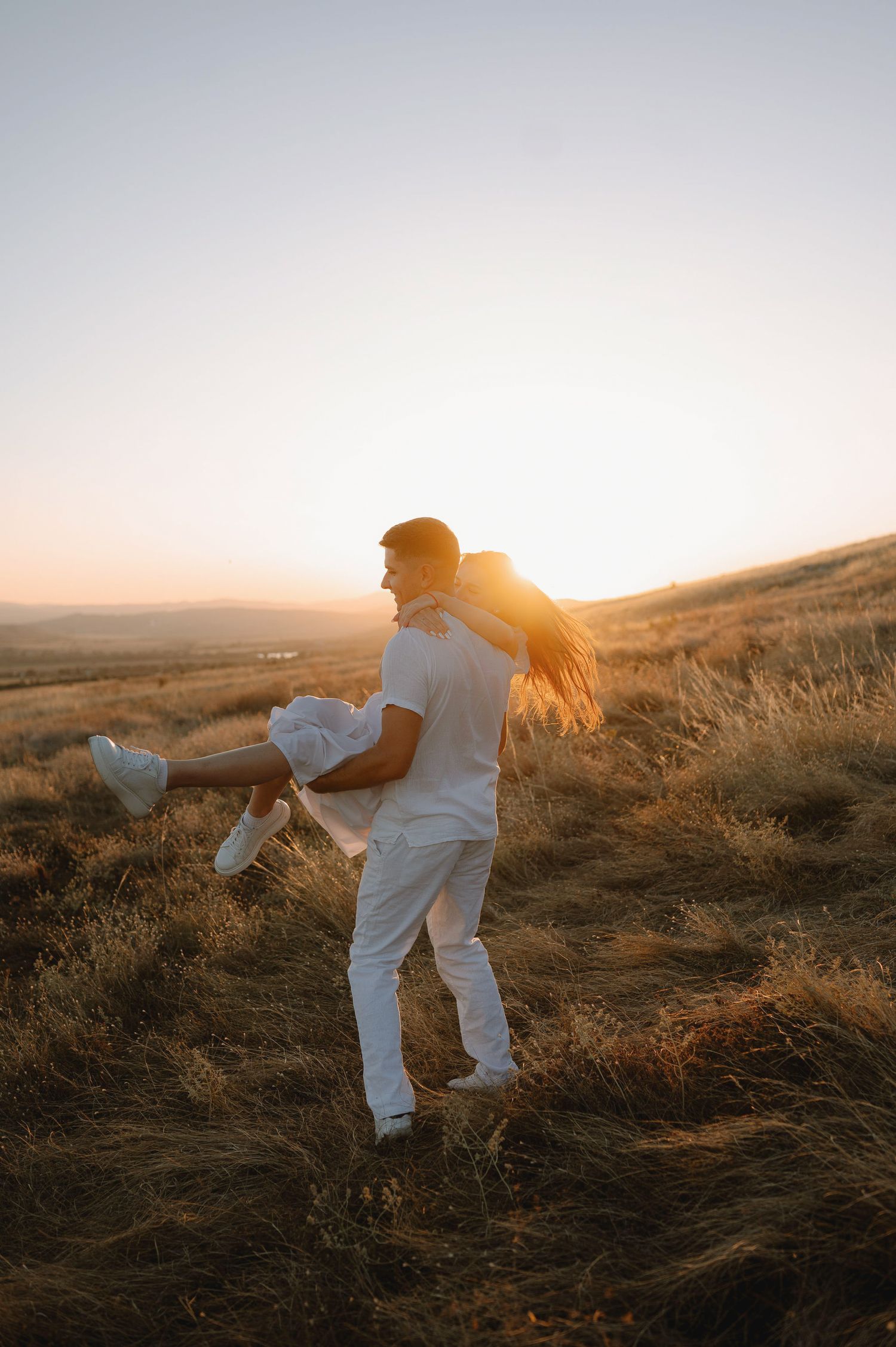 Couple in white outfits dancing together on a hillside during golden sunset hour.