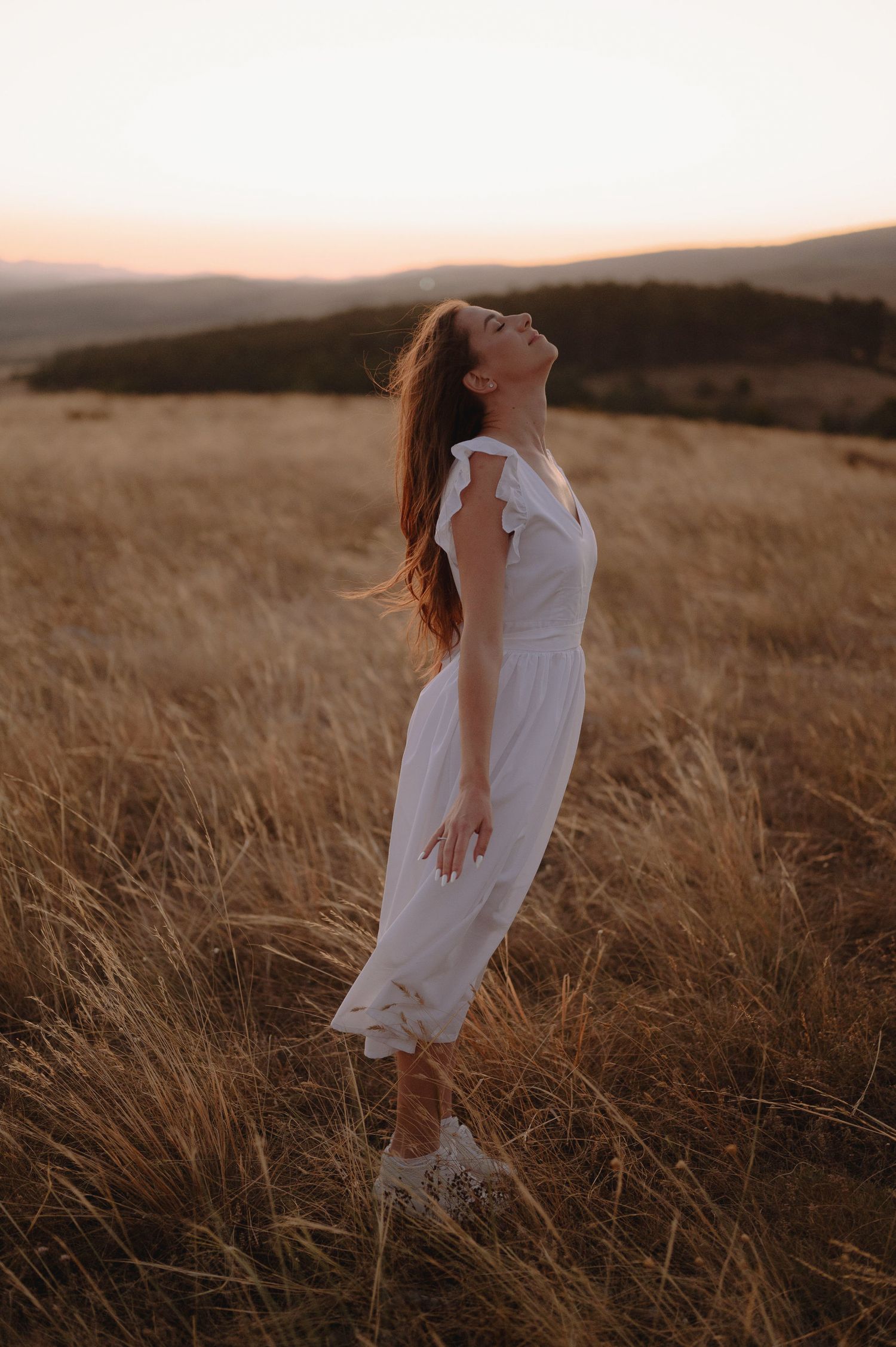 A person in a flowing white dress dances gracefully through a golden field at sunset in the mountains.