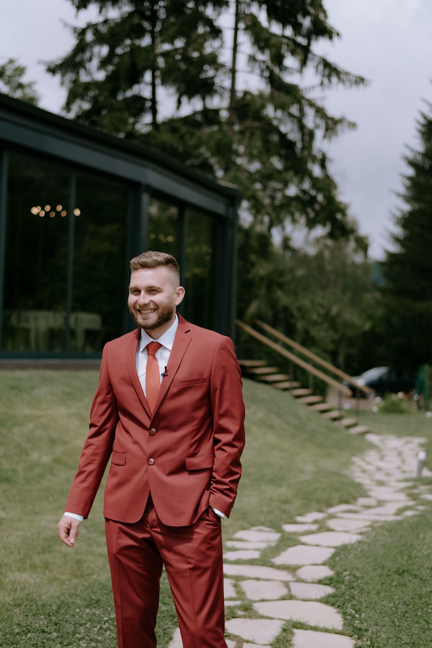 A person wearing a rust-colored suit walks along a stone pathway surrounded by green grass and trees.
