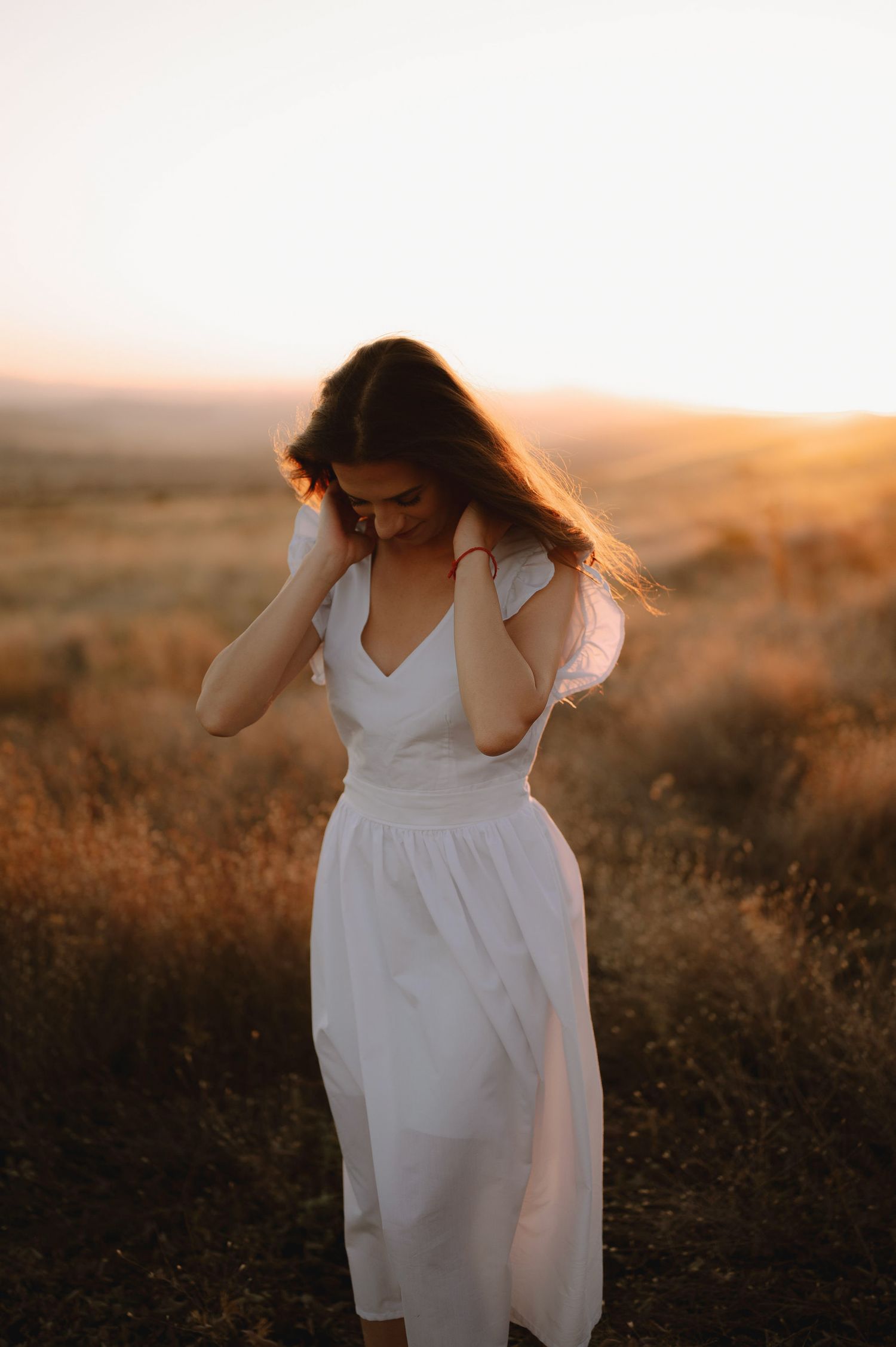 A figure in a flowing white dress stands in a golden field at sunset, their hair blowing in the wind.