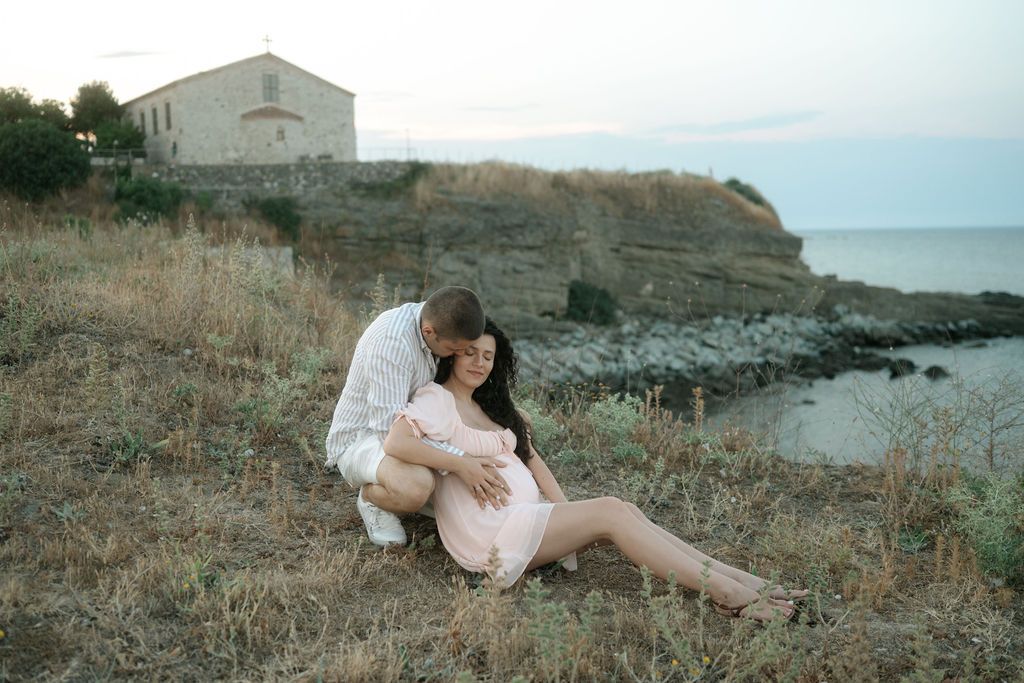 A couple shares an intimate moment on a coastal cliff with a white building and ocean in the background at sunset.