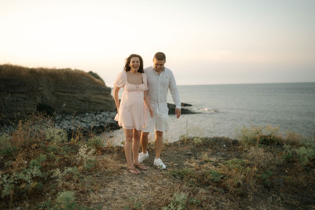 Two people in white dresses standing on a rocky coastal overlook at sunset.
