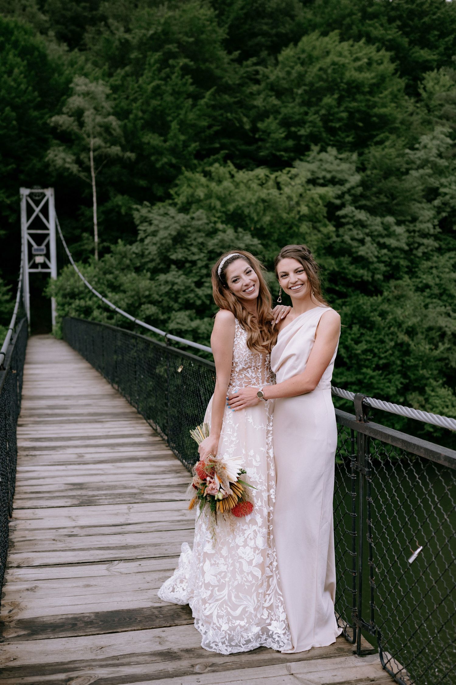 Two people in white lace dresses pose together on a wooden suspension bridge surrounded by lush green forest.