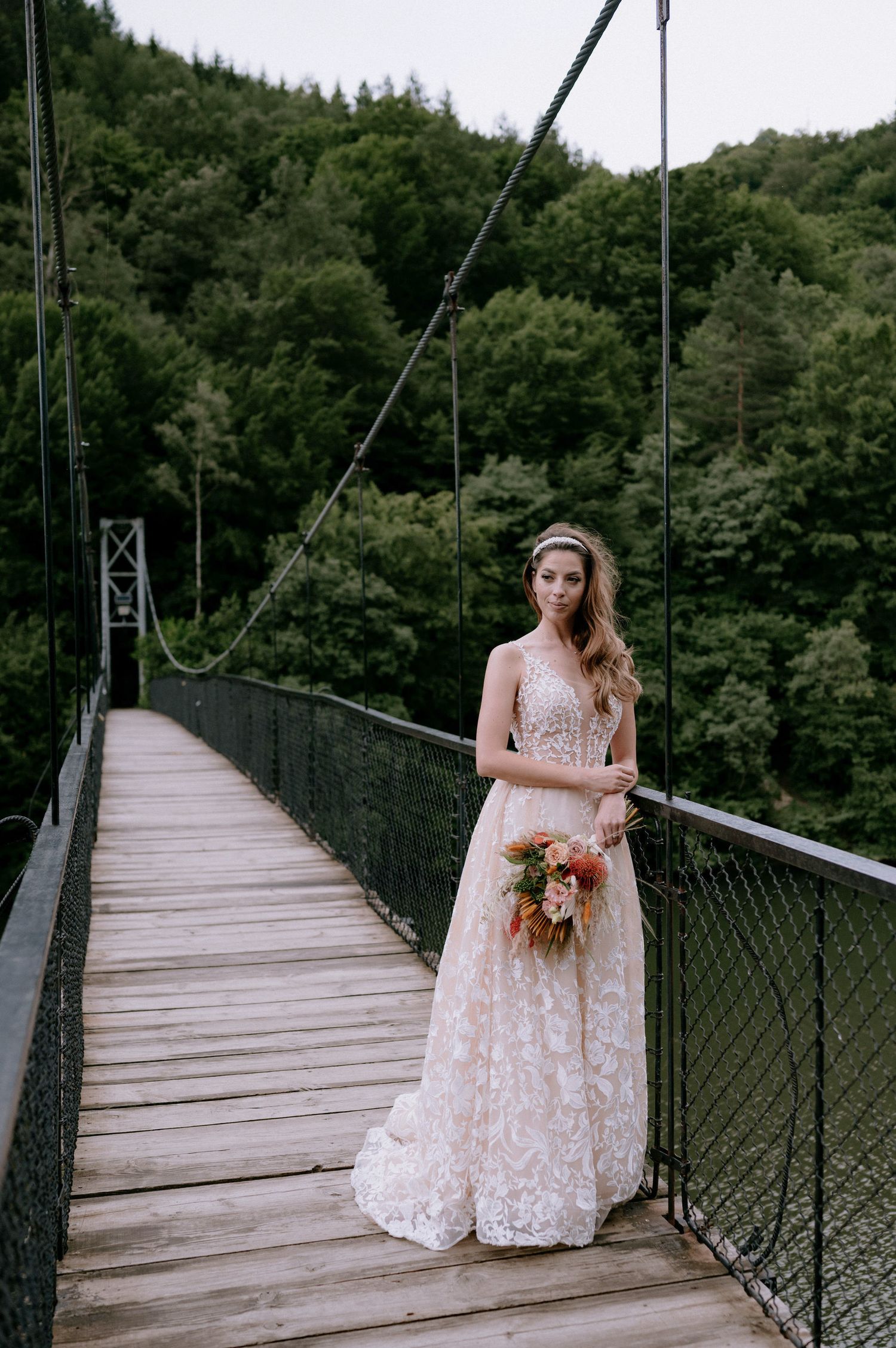 A figure in a white lace wedding dress stands on a rustic wooden suspension bridge surrounded by lush forest scenery.
