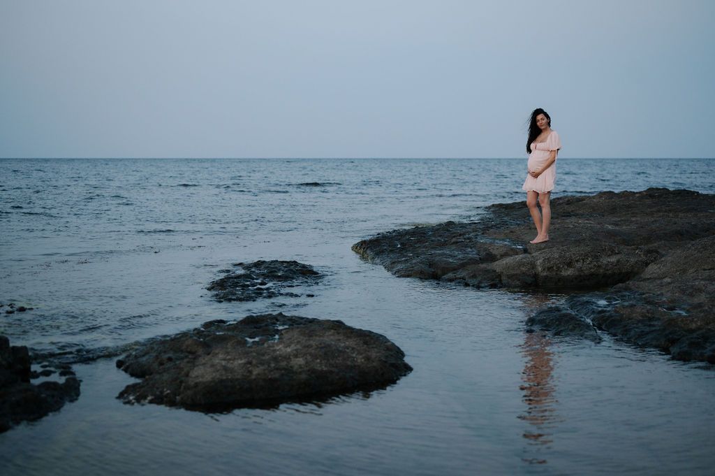 A figure stands on dark rocky coastal formations at the edge of a calm ocean during low tide.