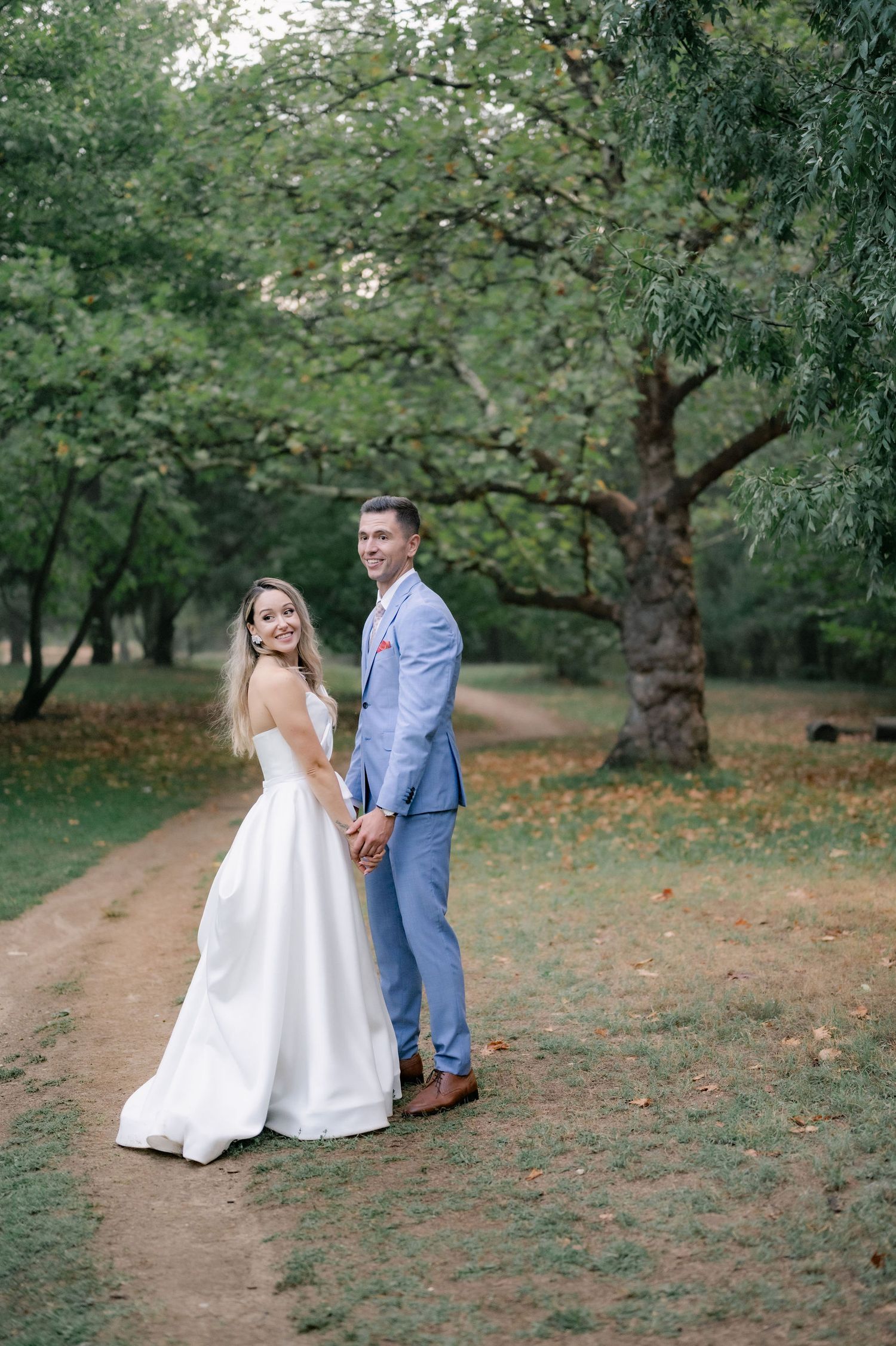 A romantic wedding couple walks along a tree-lined path in an orchard during an outdoor autumn photo session.