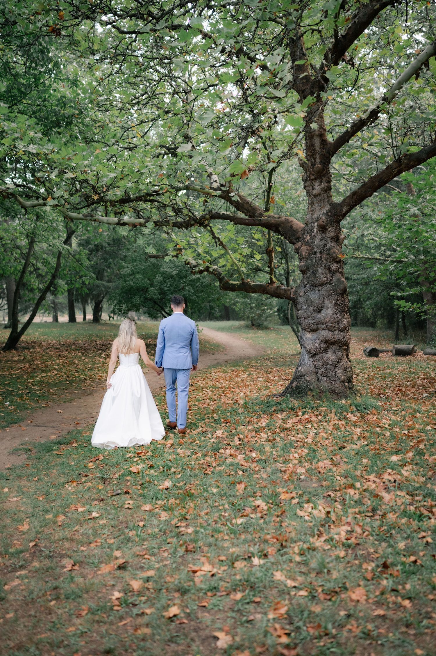 A couple walks together on a winding autumn path through a park with fallen leaves beneath old trees.