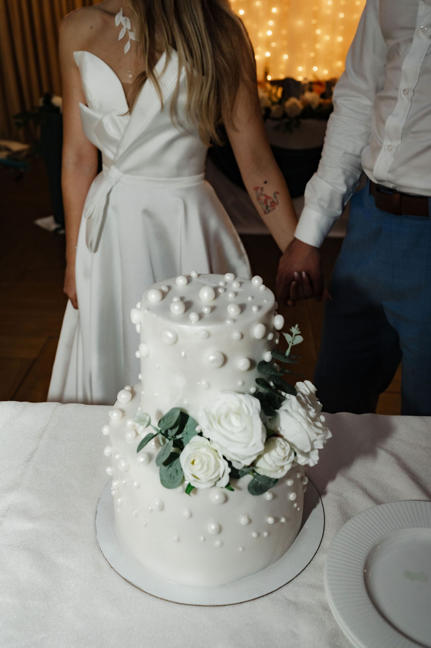 Elegant white wedding cake decorated with white roses, pearls, and eucalyptus on a white tablecloth.