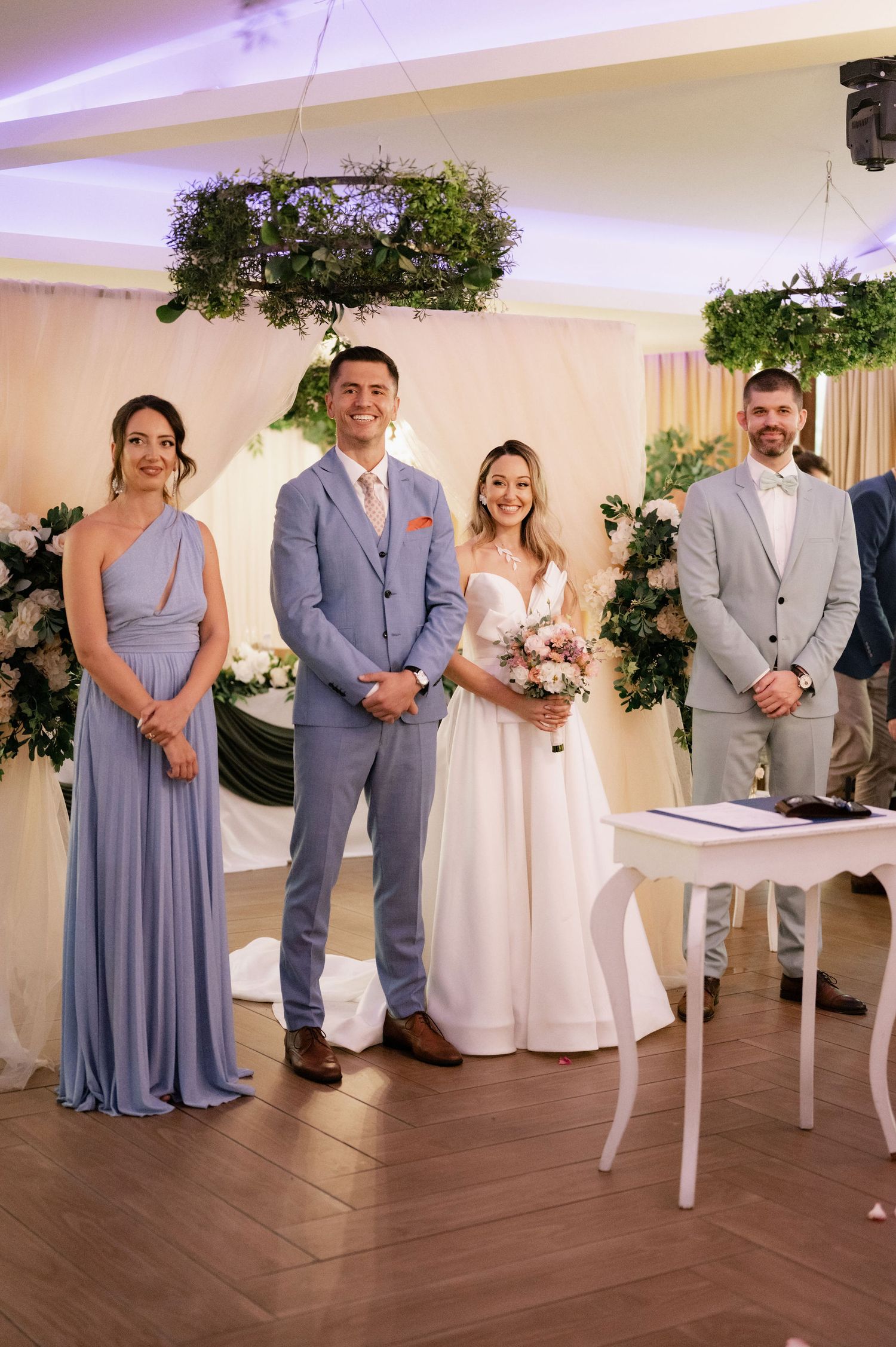 Wedding party members stand together at an indoor ceremony with hanging greenery and white decor on hardwood floors.